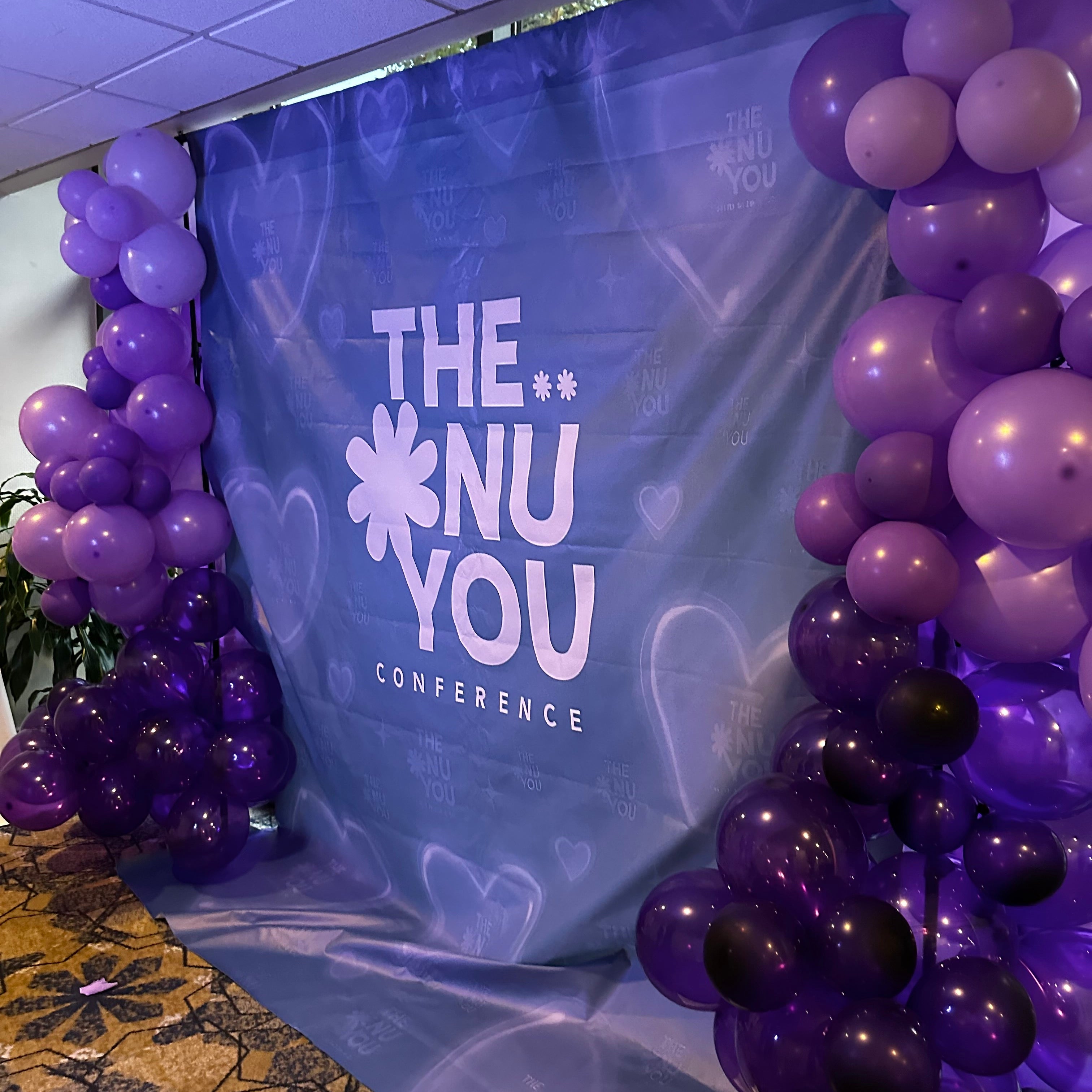 Photo booth area at the NuMomish The Nu You Conference framed with purple balloons, the event featured The Dappled Wood as a vendor showcasing handmade jewelry.