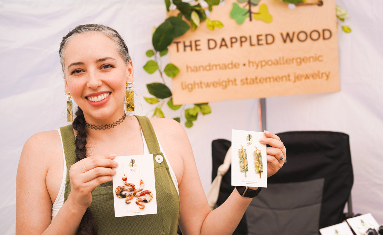 The founder of The Dappled Wood smiling in green overalls, holding pairs of handmade polymer clay earrings with The Dappled Wood sign partially visible in the background.