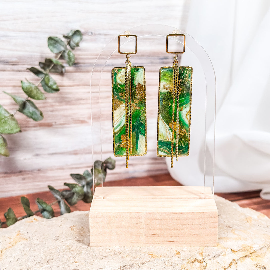 The Dappled Wood's Athena green and gold statement earrings from the Gilded Grove Collection, featuring rectangular faux agate panels with gold chains, displayed on a clear stand with a wooden base and eucalyptus styling.