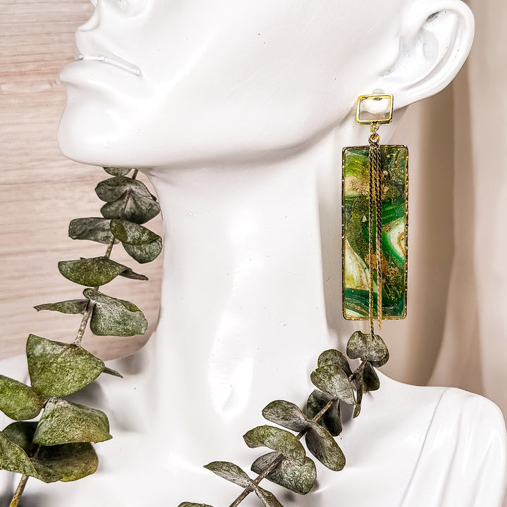 The Dappled Wood's Athena green and gold statement earrings from the Gilded Grove Collection, featuring a rectangular faux agate design with cascading gold chains, displayed on a white mannequin with eucalyptus styling.