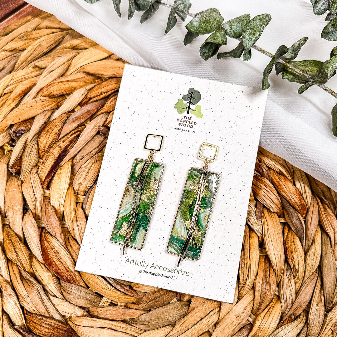 The Dappled Wood's Athena green and gold statement earrings from the Gilded Grove Collection, featuring rectangular faux agate marbled panels with gold chain details, displayed on a branded card with a woven background and eucalyptus sprigs.