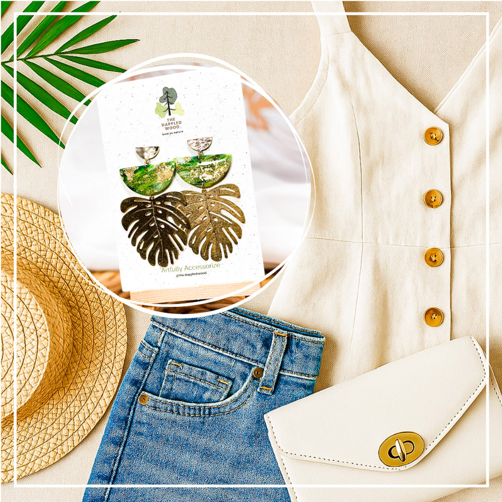 The Dappled Wood's Isolde green and gold statement earrings with leaves from the Gilded Grove Collection, styled with a casual summer outfit featuring a linen top, denim, straw hat, and clutch.