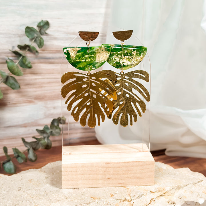 The Dappled Wood's Isolde green and gold leaf statement earrings from the Gilded Grove Collection, shown on a clear acrylic display stand with a wood base and eucalyptus accents.