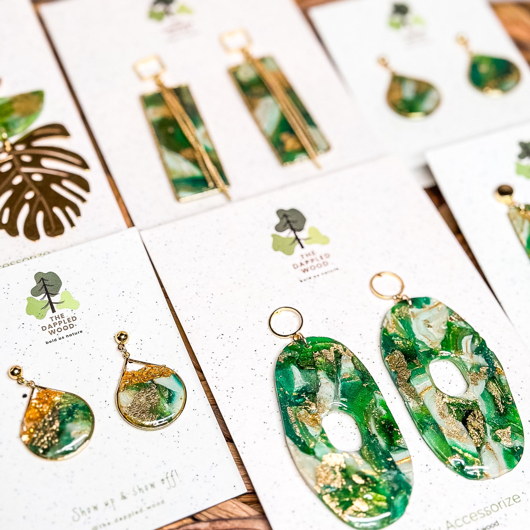 Close-up flat lay of The Dappled Wood Gilded Grove Collection Spring 2025 earrings, featuring Chloe and Elowen designs in vibrant greens accented with gold leaf, displayed on branded earring cards, highlighting nature-inspired lightweight hypoallergenic jewelry.