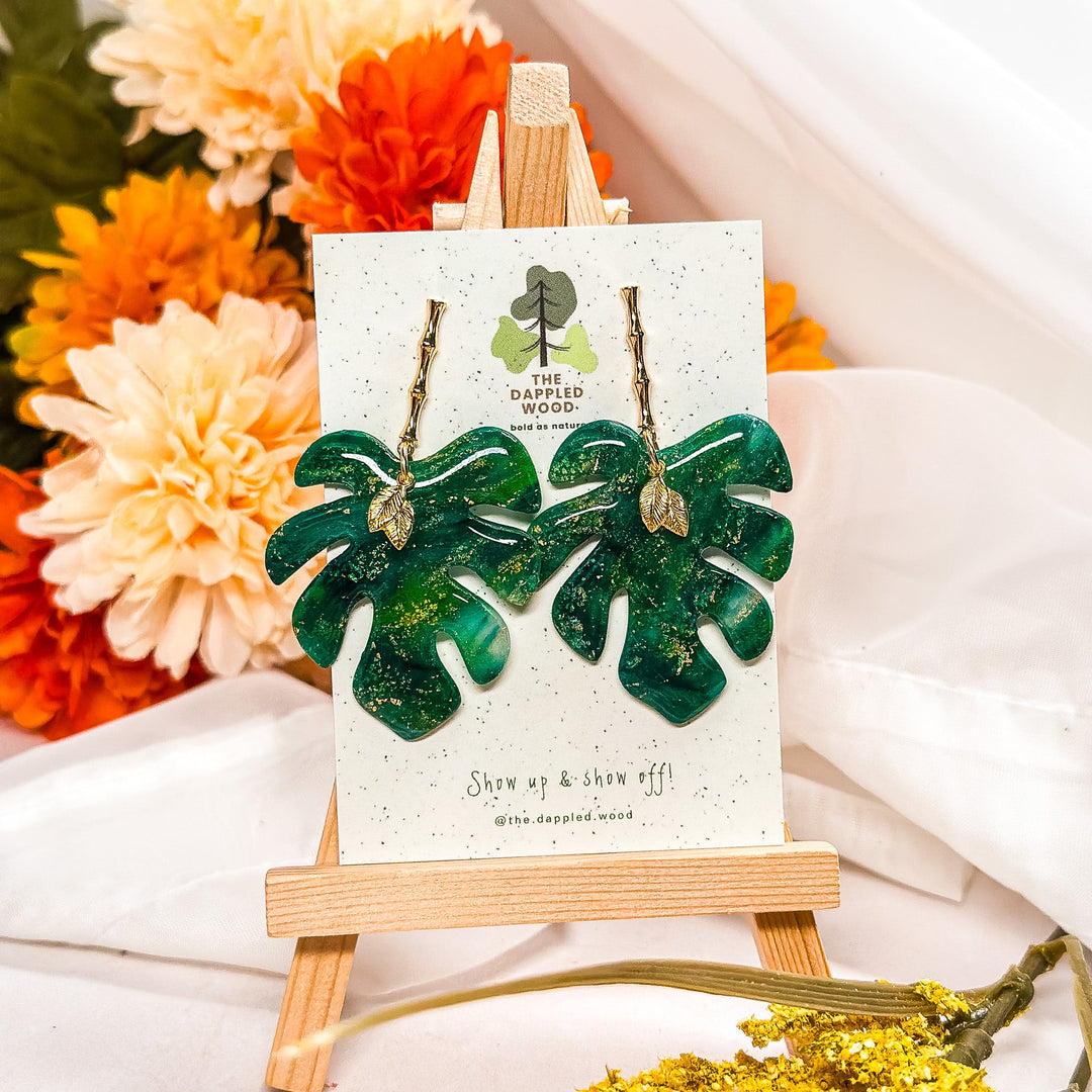 Sparkly green monstera leaf polymer clay earrings with gold hooks from The Dappled Wood’s All Styles collection, displayed on an easel with flowers.