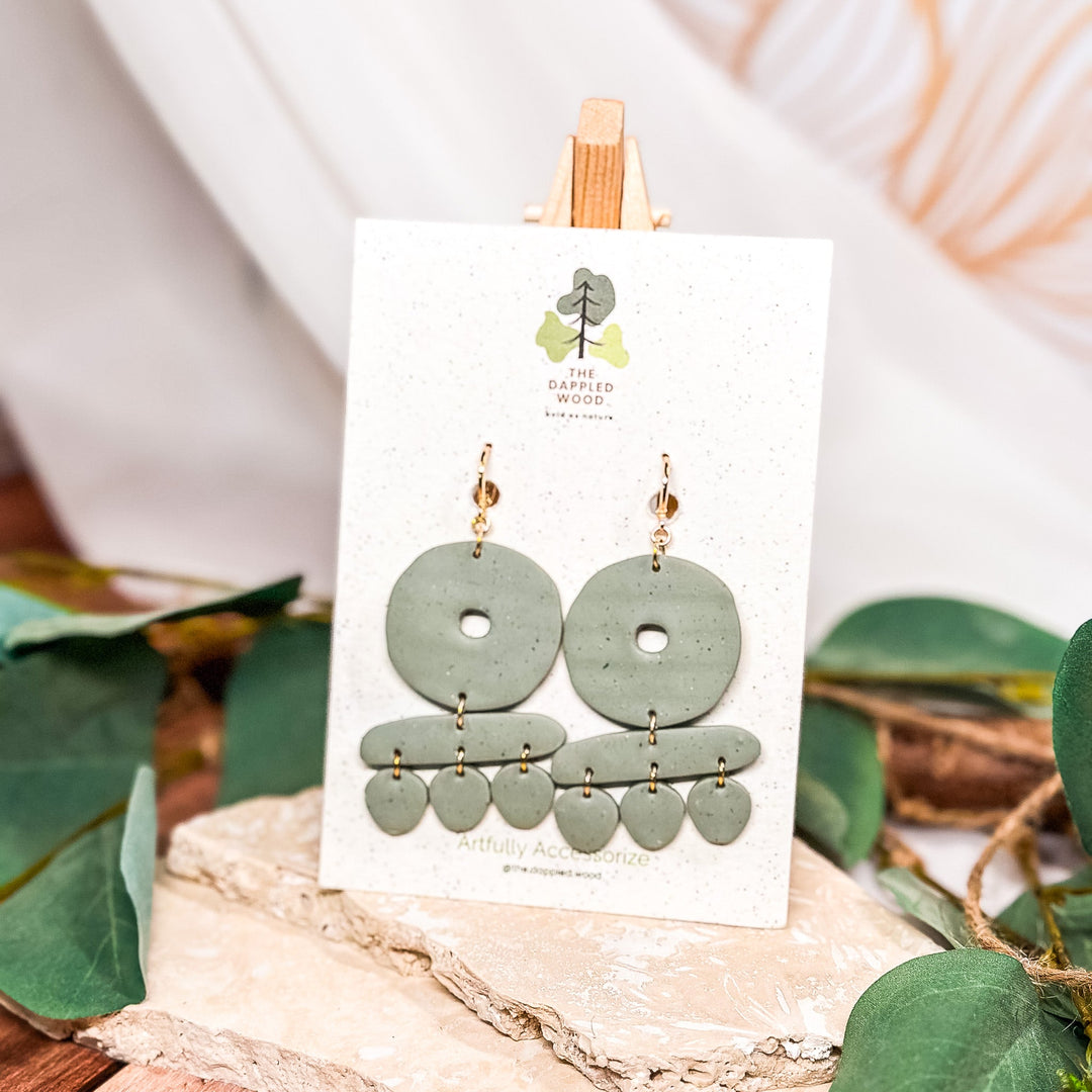 Sage green polymer clay dangle earrings from The Dappled Wood’s New Arrivals collection, displayed on a stand with natural foliage accents.