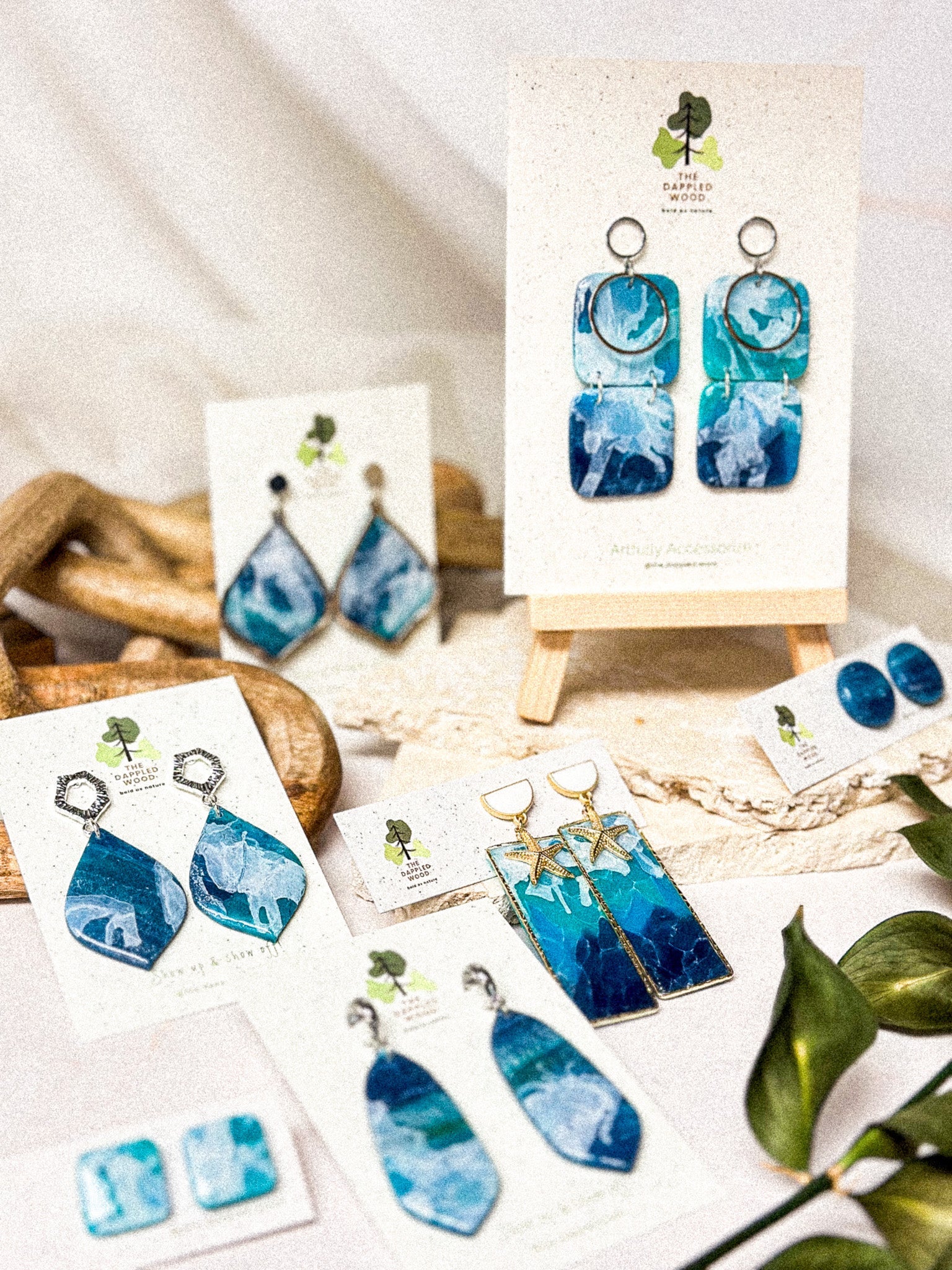 Ocean-inspired blue polymer clay earrings from The Dappled Wood’s Surf & Sun Summer 2024 Earring Collection, displayed on stands with beachy accents.