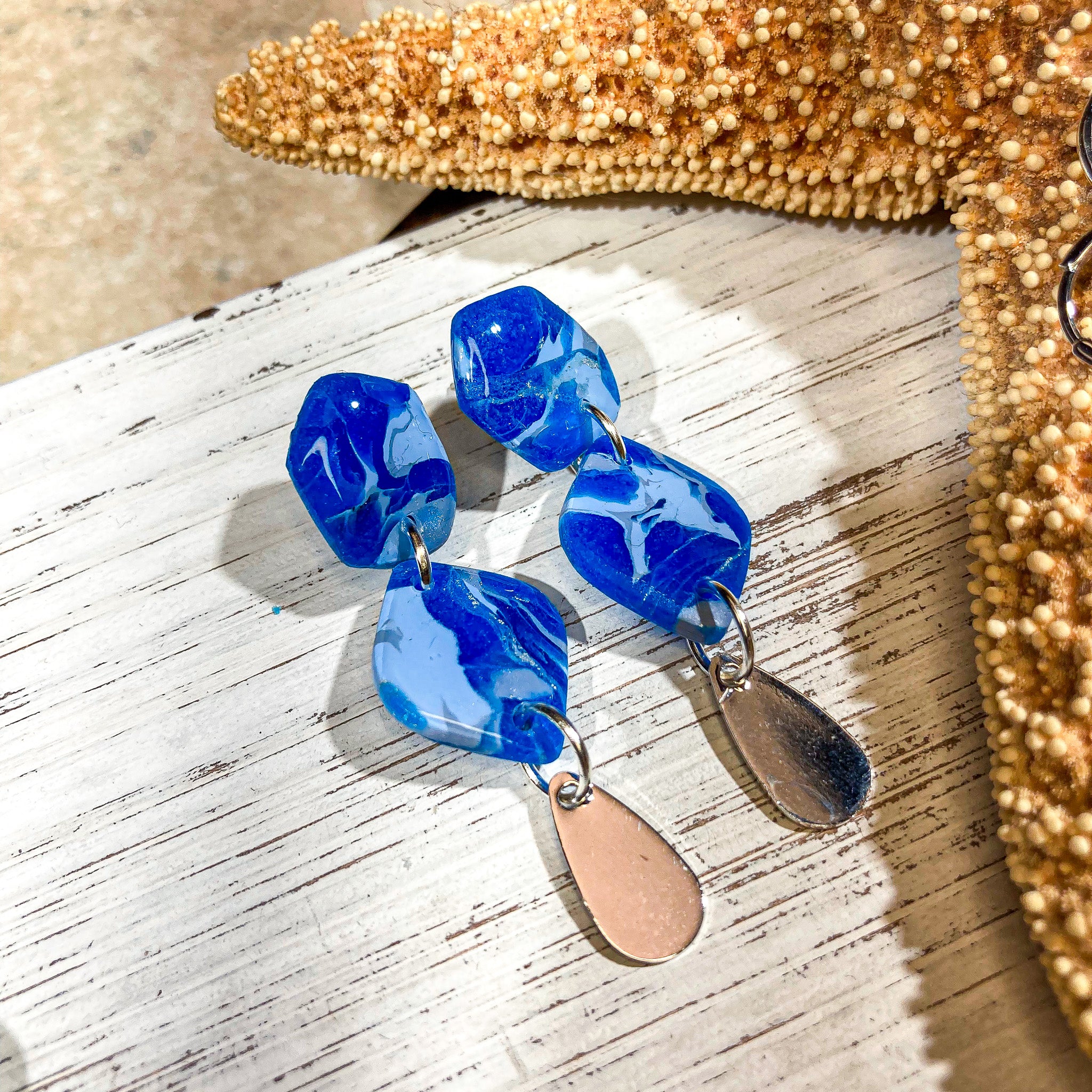Handcrafted polymer clay earrings in marbled cornflower and sapphire blue with a silver teardrop charm, displayed on a white wooden surface with a starfish decor in the background.