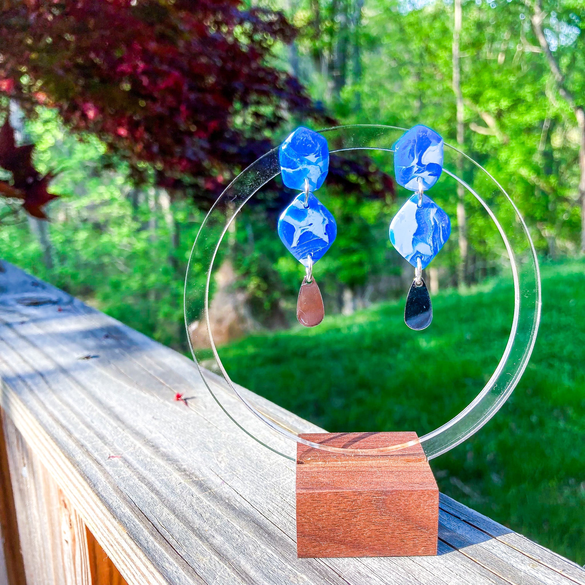 Handcrafted polymer clay earrings in marbled cornflower and sapphire blue with a silver teardrop charm, displayed on a clear acrylic circle with a warm wood base which is sitting on a fence with lush woods in the background.