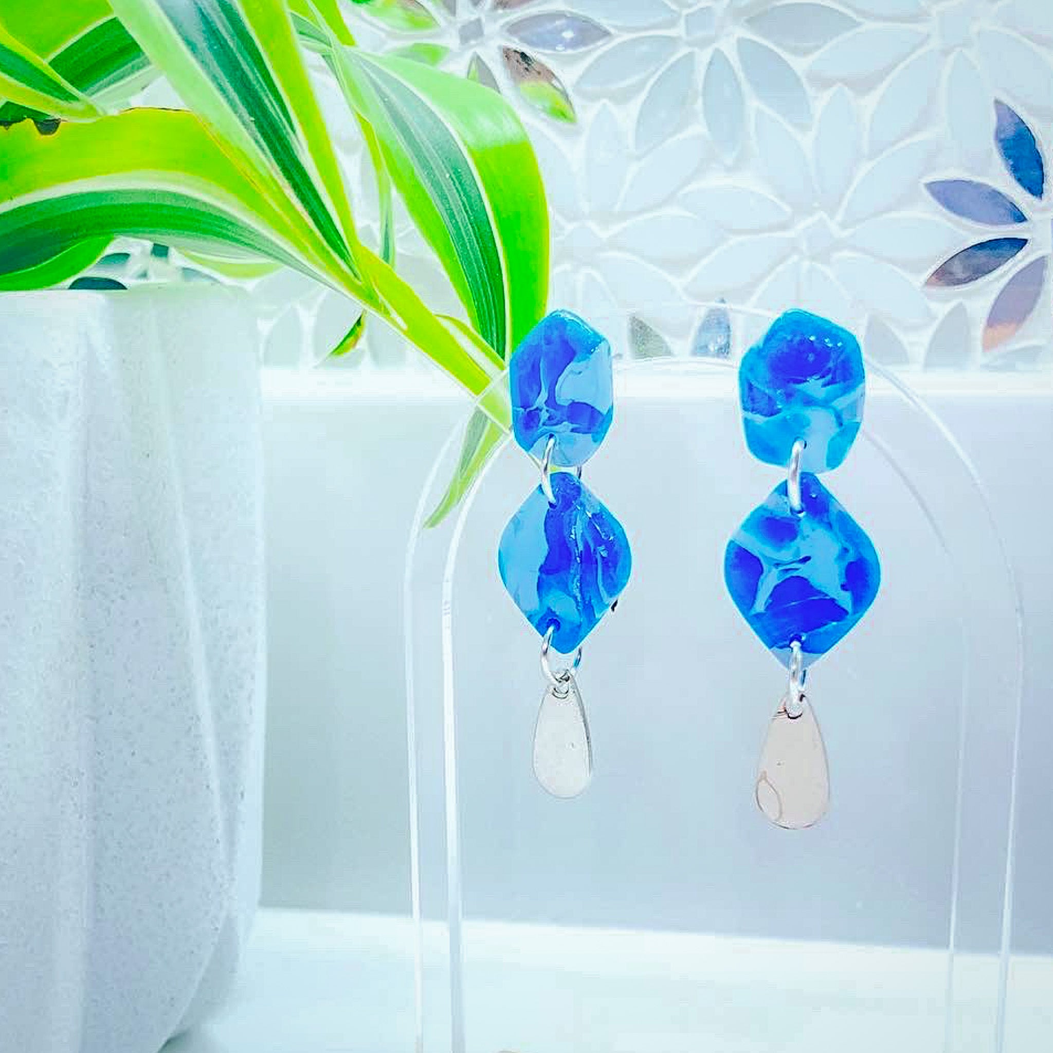 Handcrafted polymer clay earrings in marbled cornflower and sapphire blue with a silver teardrop charm, displayed on a clear acrylic arch with a bright green plant in a white geometric pot in the background.