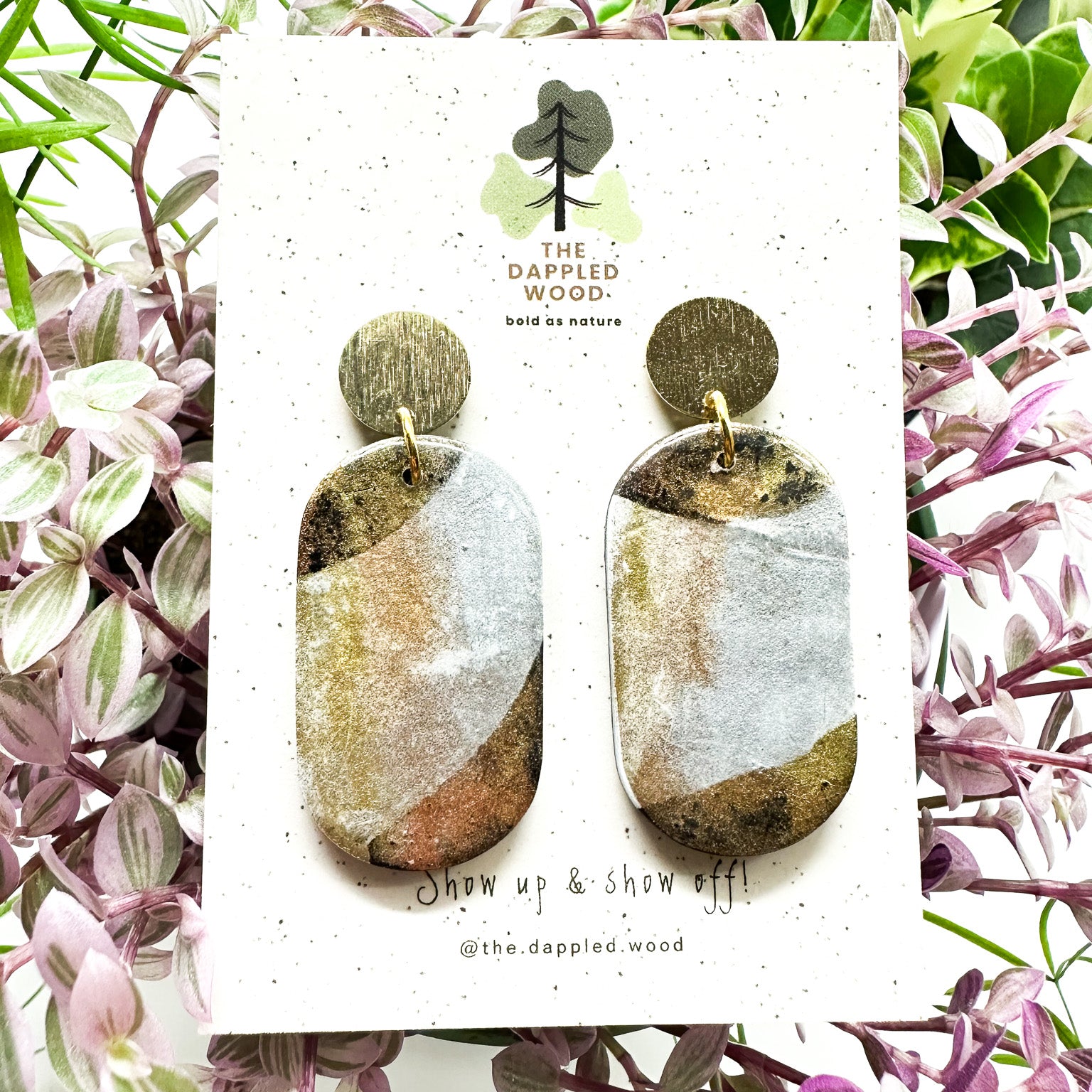 Oval-shaped polymer clay earrings with gradient earthy tones set on a white card, surrounded by delicate purple and green plants. Card bears 'The Dappled Wood' logo and the phrase 'Show up & show off!'.