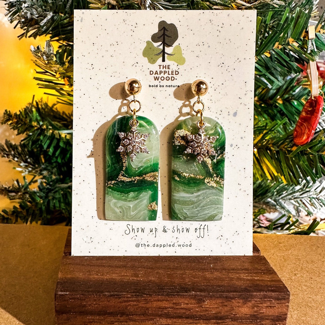 Handcrafted polymer clay earrings with a faux jade agate design displayed on a cardboard holder, embellished with a gold-colored snowflake charm. The earrings are presented against a festive backdrop with a Christmas tree, emphasizing their holiday theme. The holder features the brand name 'The Dappled Wood' with the tagline 'Show up & show off!' and the earrings are resting on a dark wooden stand.