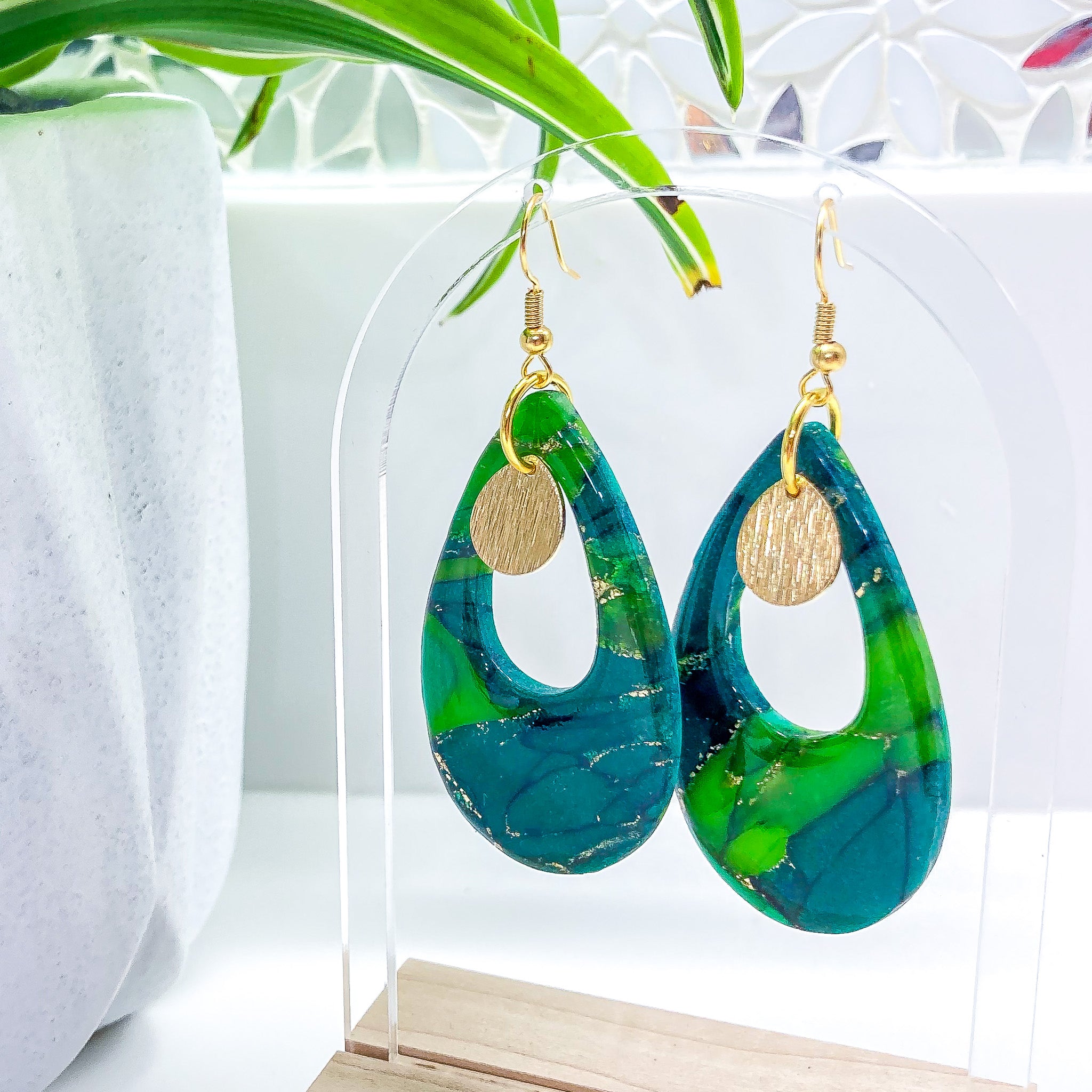 Elegant teardrop-shaped polymer clay earrings featuring a marbled emerald and teal design with a brushed gold disc, showcased against a modern white display and green foliage background.