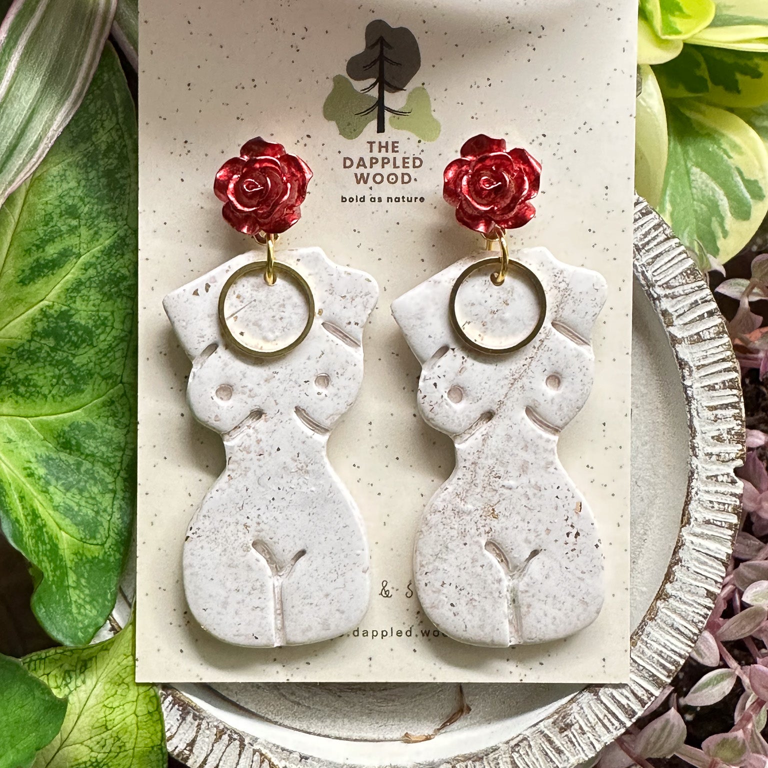 Elegant white polymer clay earrings with gold specks in the shape of a natural, curvy woman with a gold circle pendant, hanging from radiant red and gold rose posts. Earrings are showcased on a branded 'The Dappled Wood' card surrounded by lush greenery.