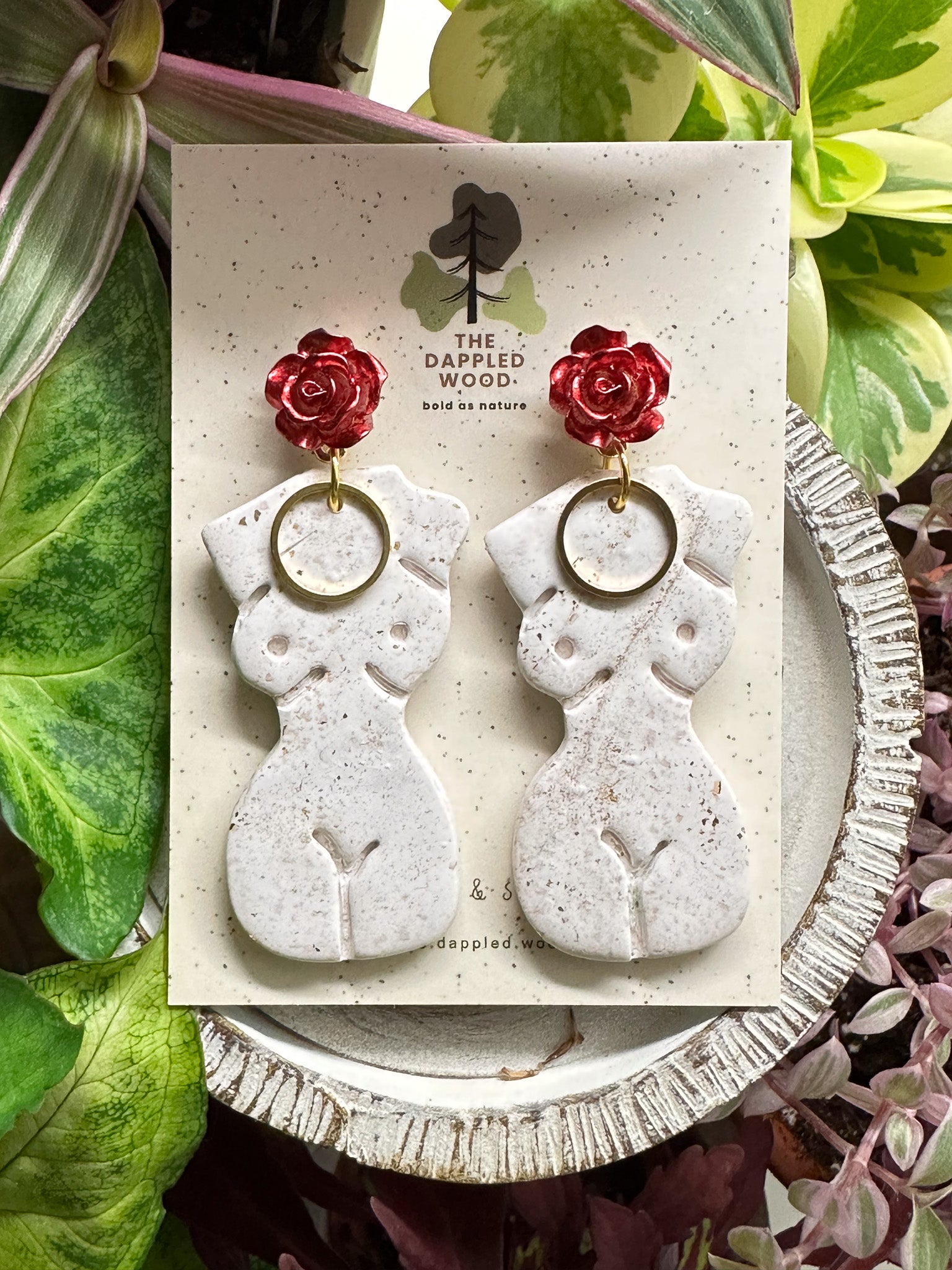 Elegant white polymer clay earrings with gold specks in the shape of a natural, curvy woman with a gold circle pendant, hanging from radiant red and gold rose posts. Earrings are showcased on a branded 'The Dappled Wood' card surrounded by lush greenery.