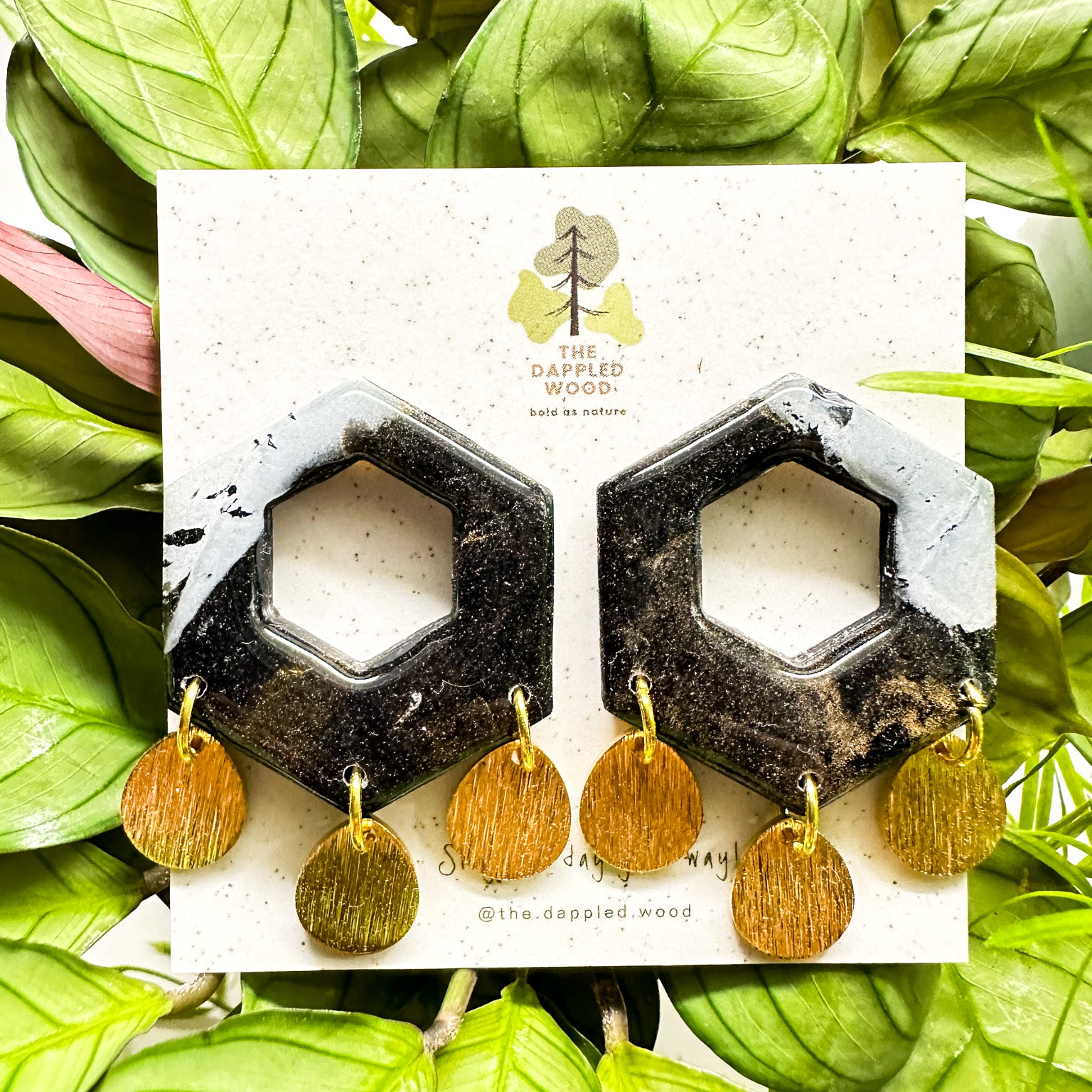 Black and glittered hexagonal-shaped polymer clay earrings complemented with small round gold textured dangles, displayed on a white card surrounded by vibrant green leaves. Card features 'The Dappled Wood' logo.