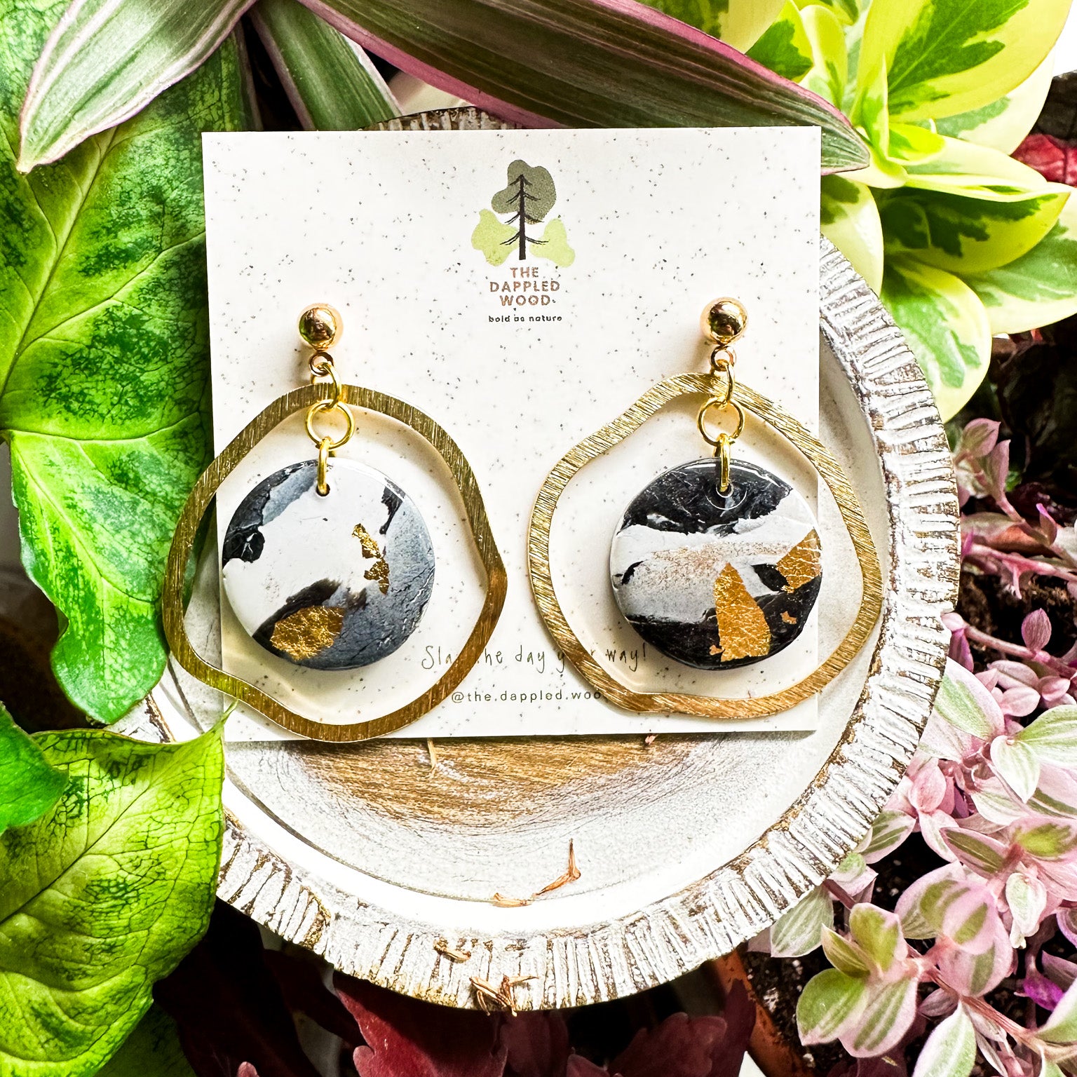 Handcrafted black and white marbled circular polymer clay earrings with gold accents displayed on a speckled card labeled 'The Dappled Wood' surrounded by vibrant indoor plants in shades of green, pink, and purple.