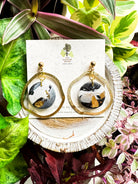 Handcrafted black and white marbled circular polymer clay earrings with gold accents displayed on a speckled card labeled 'The Dappled Wood' surrounded by vibrant indoor plants in shades of green, pink, and purple.