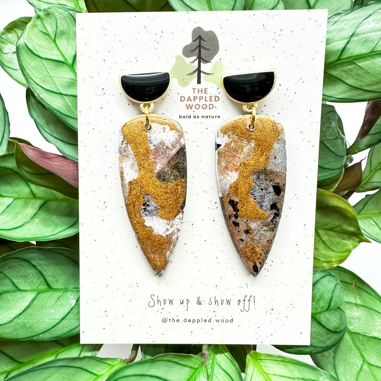 Dagger-shaped metallic coated polymer clay earrings with black and gold half moon posts are displayed on a speckled card with The Dappled Wood branding, midst vibrant green leaves.