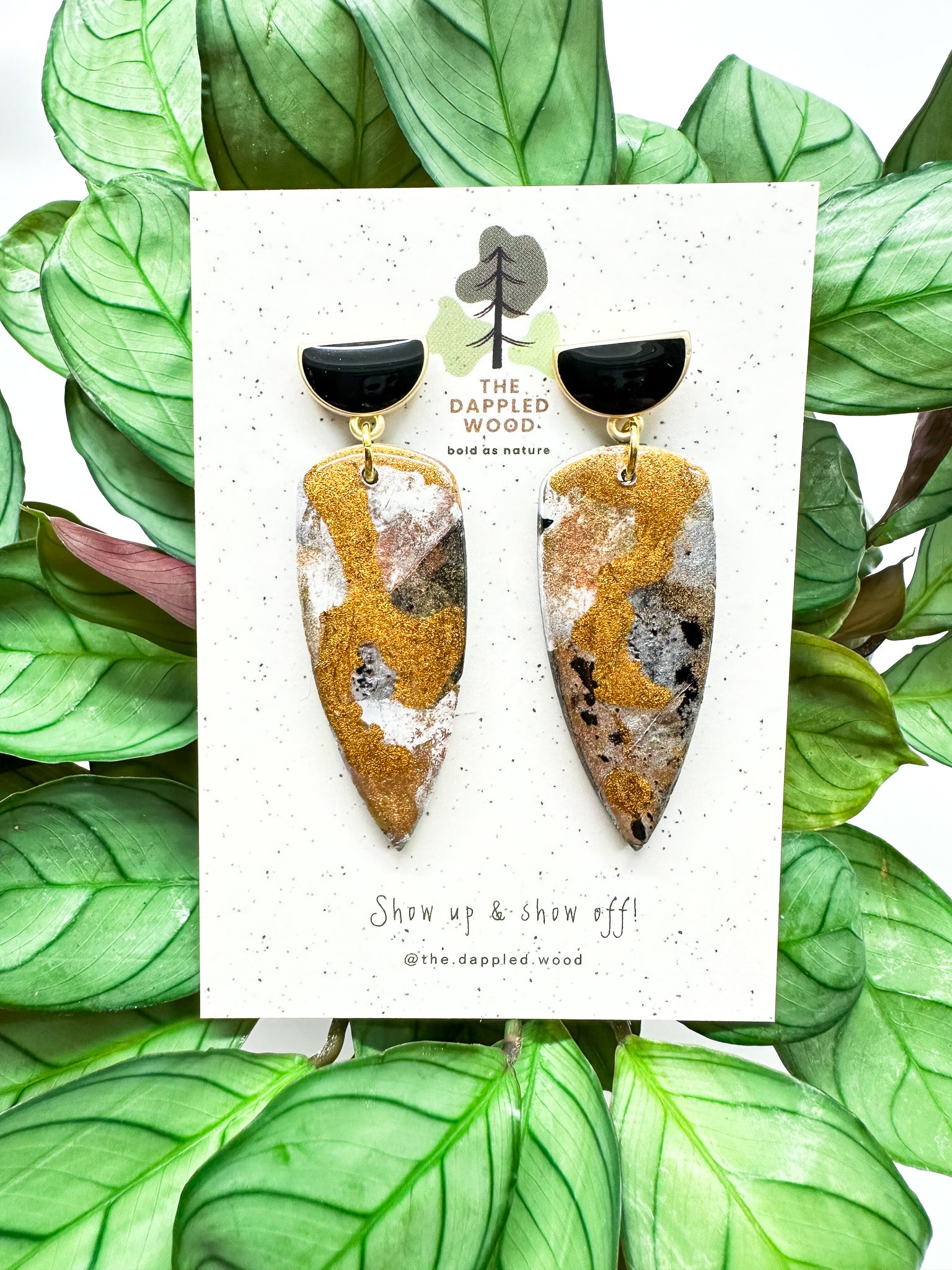 Dagger-shaped metallic coated polymer clay earrings with black and gold half moon posts are displayed on a speckled card with The Dappled Wood branding, midst vibrant green leaves.