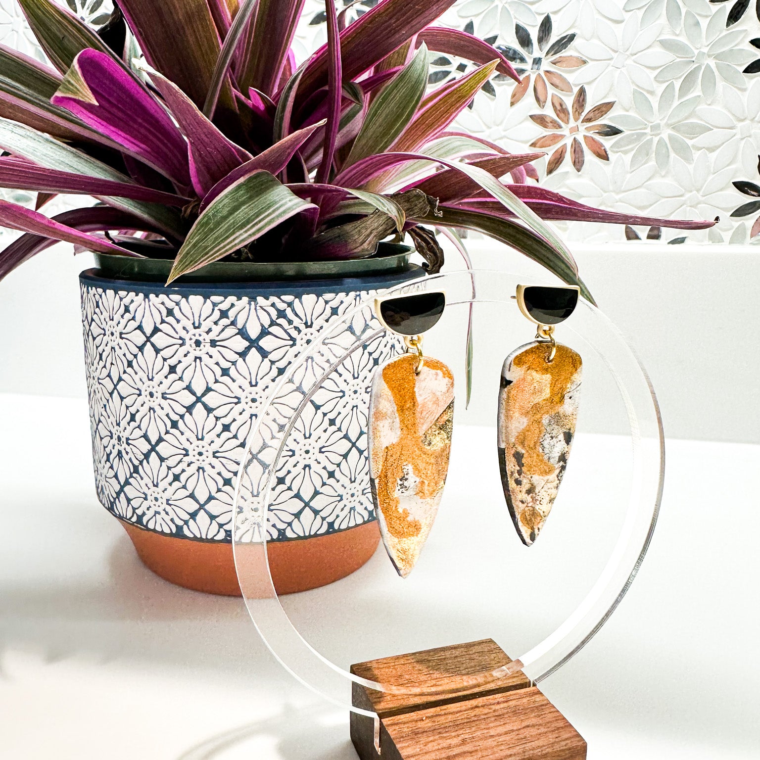 Elegant metallic polymer clay earrings with gold and black half moon accents dangle a clear circular display with a wooden block base, set against a mosaic backdrop with a rich purple-leafed plant in a blue and white patterned pot.