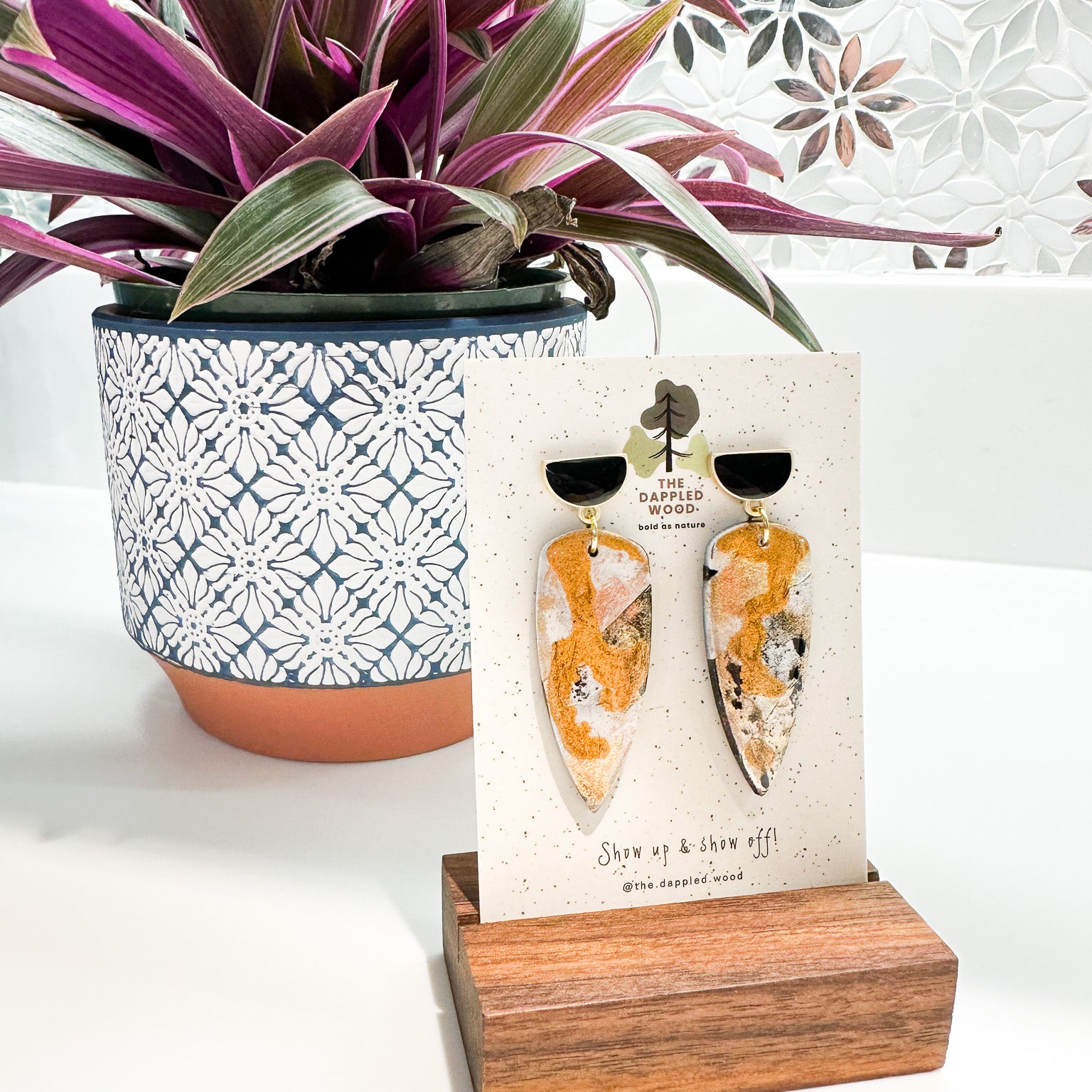 Handcrafted dagger shaped polymer clay earrings with gold flecks displayed on a branded card by @the.dappled.wood, placed against a decorative mosaic backdrop with a vibrant purple-leafed plant in a patterned pot.