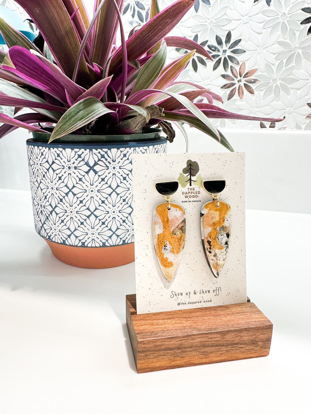 Handcrafted dagger shaped polymer clay earrings with gold flecks displayed on a branded card by @the.dappled.wood, placed against a decorative mosaic backdrop with a vibrant purple-leafed plant in a patterned pot.