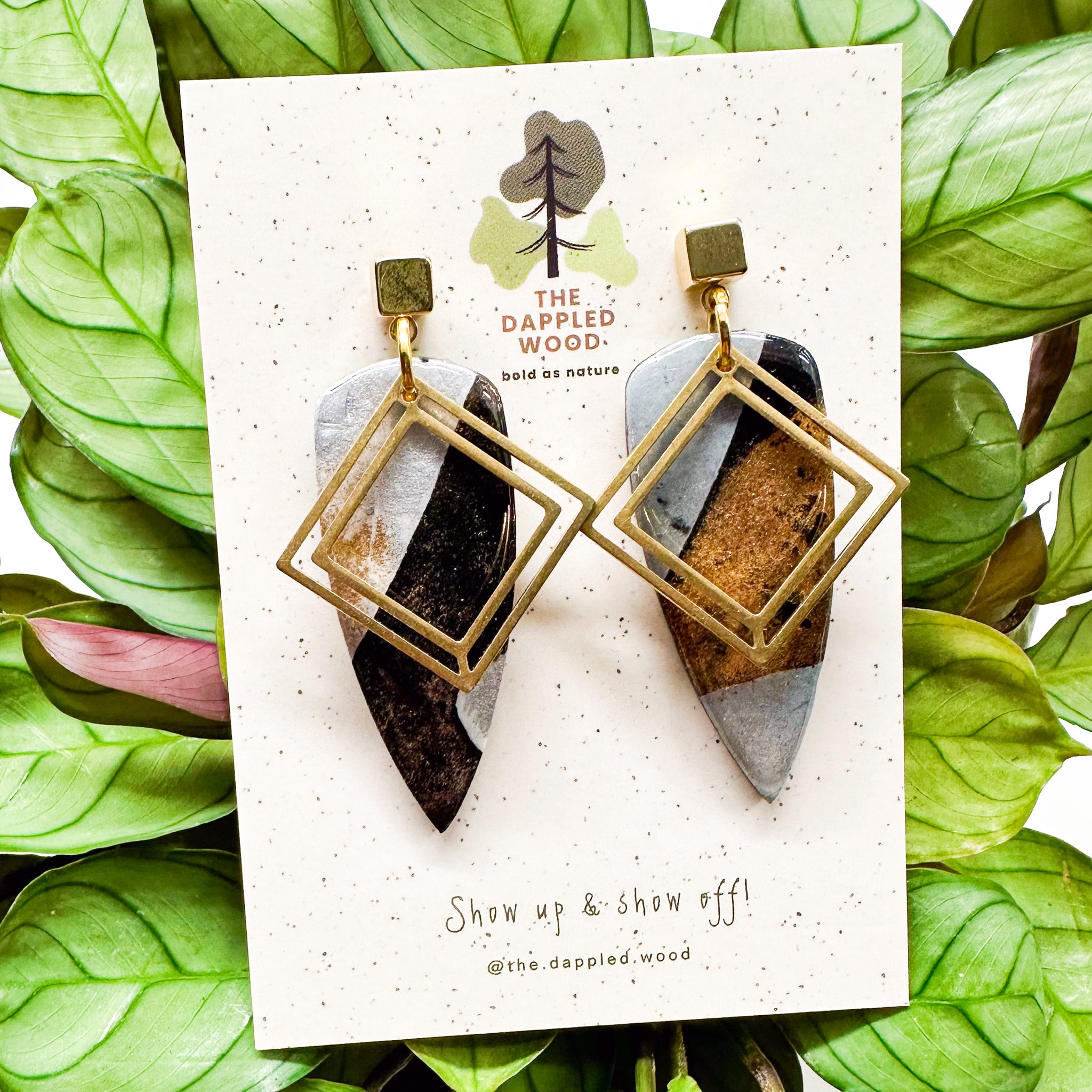 Unique geometric polymer clay earrings with gold frames and earthy toned insets, hanging on a white display card with green foliage in the backdrop. The card showcases 'The Dappled Wood' logo and the slogan 'Show up & show off!'.