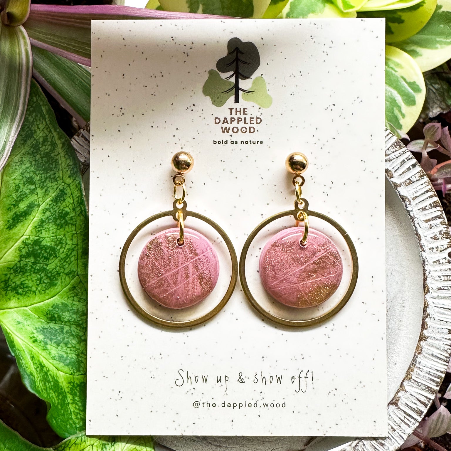 Handcrafted 'Michele' polymer clay earrings featuring shimmering pink circular pendants with gold hoops, displayed on a speckled white card with nature-inspired 'The Dappled Wood' branding, complemented by a lush backdrop of green and pink foliage.