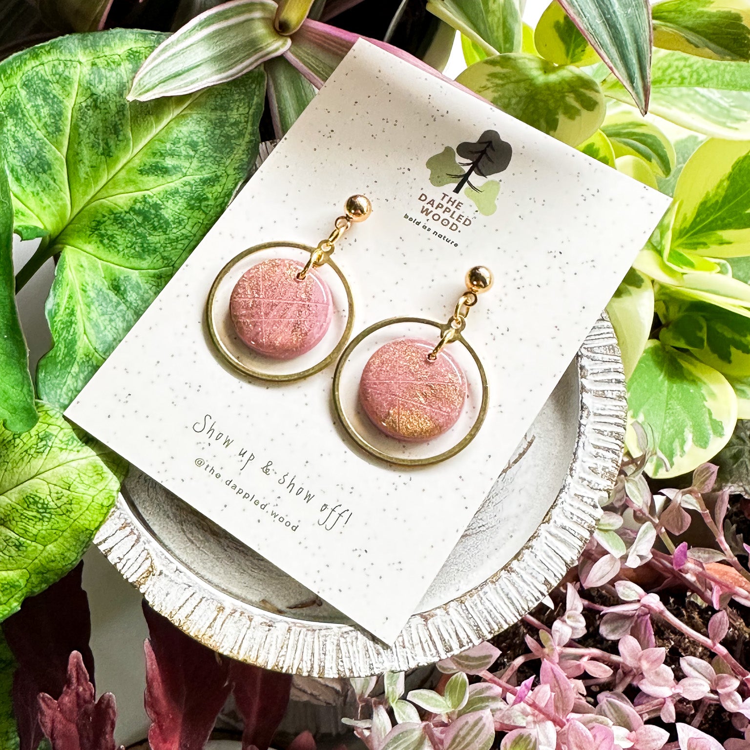 Handcrafted 'Michele' polymer clay earrings featuring shimmering pink circular pendants with gold hoops, displayed on a speckled white card with nature-inspired 'The Dappled Wood' branding, complemented by a lush backdrop of green and pink foliage.