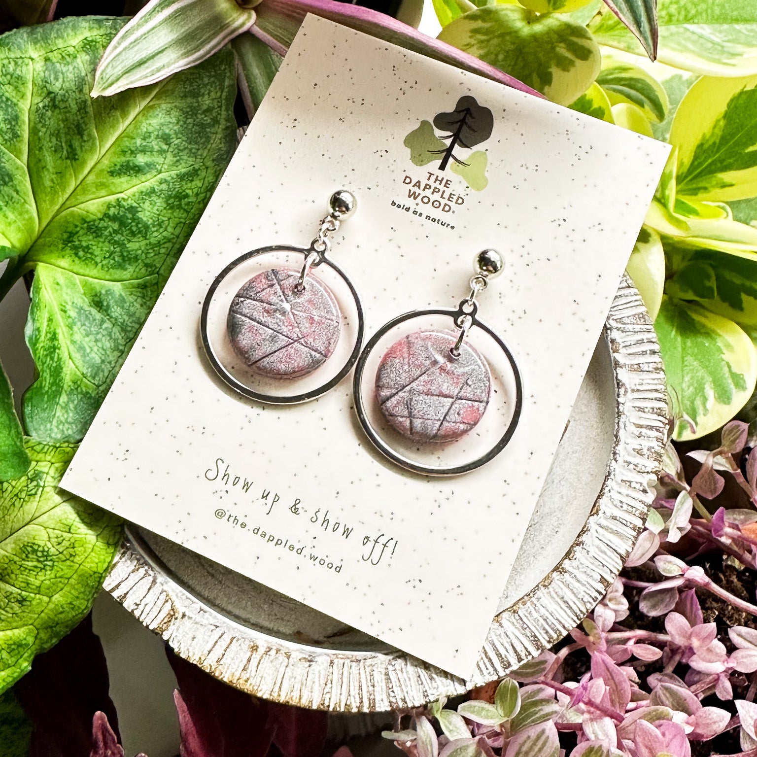Handcrafted 'Michele' polymer clay earrings featuring shimmering pink circular pendants with silver hoops, displayed on a speckled white card with nature-inspired 'The Dappled Wood' branding, complemented by a lush backdrop of green and pink foliage.