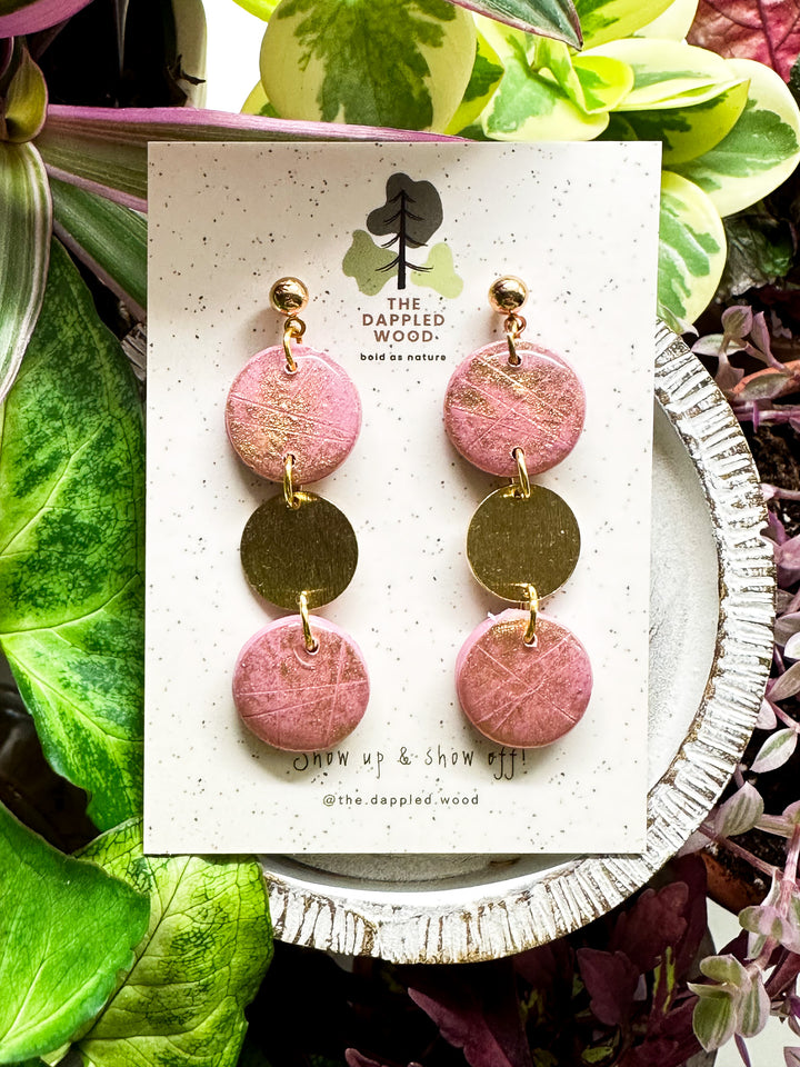 Handcrafted pink polymer clay disk earrings with gold shimmer and a textured brass connector charm, displayed on a speckled card with The Dappled Wood logo against a lush backdrop of green plants.