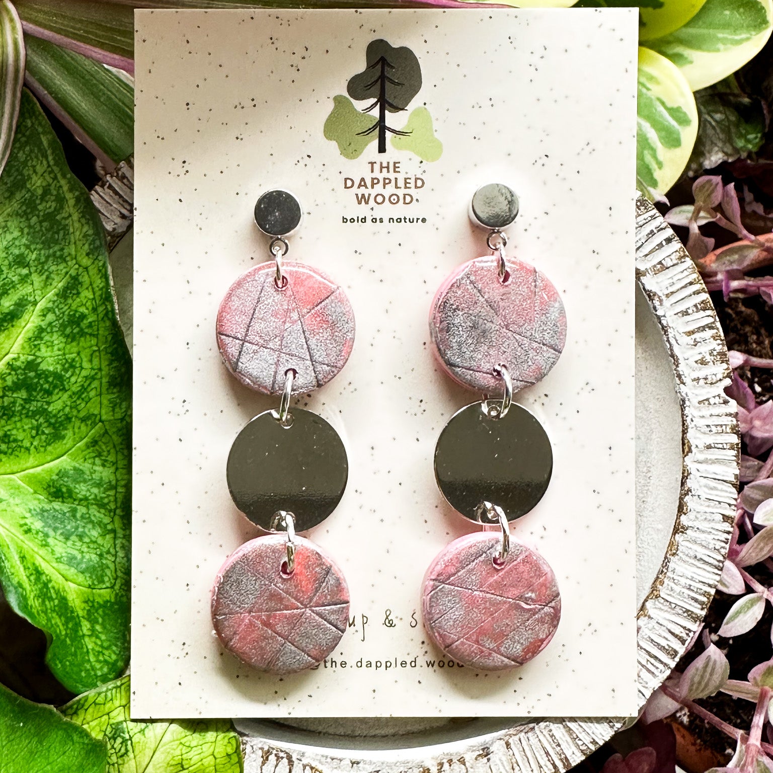 Handcrafted pink polymer clay disk earrings with silver shimmer and a smooth silver connector charm, displayed on a speckled card with The Dappled Wood logo against a lush backdrop of green plants.