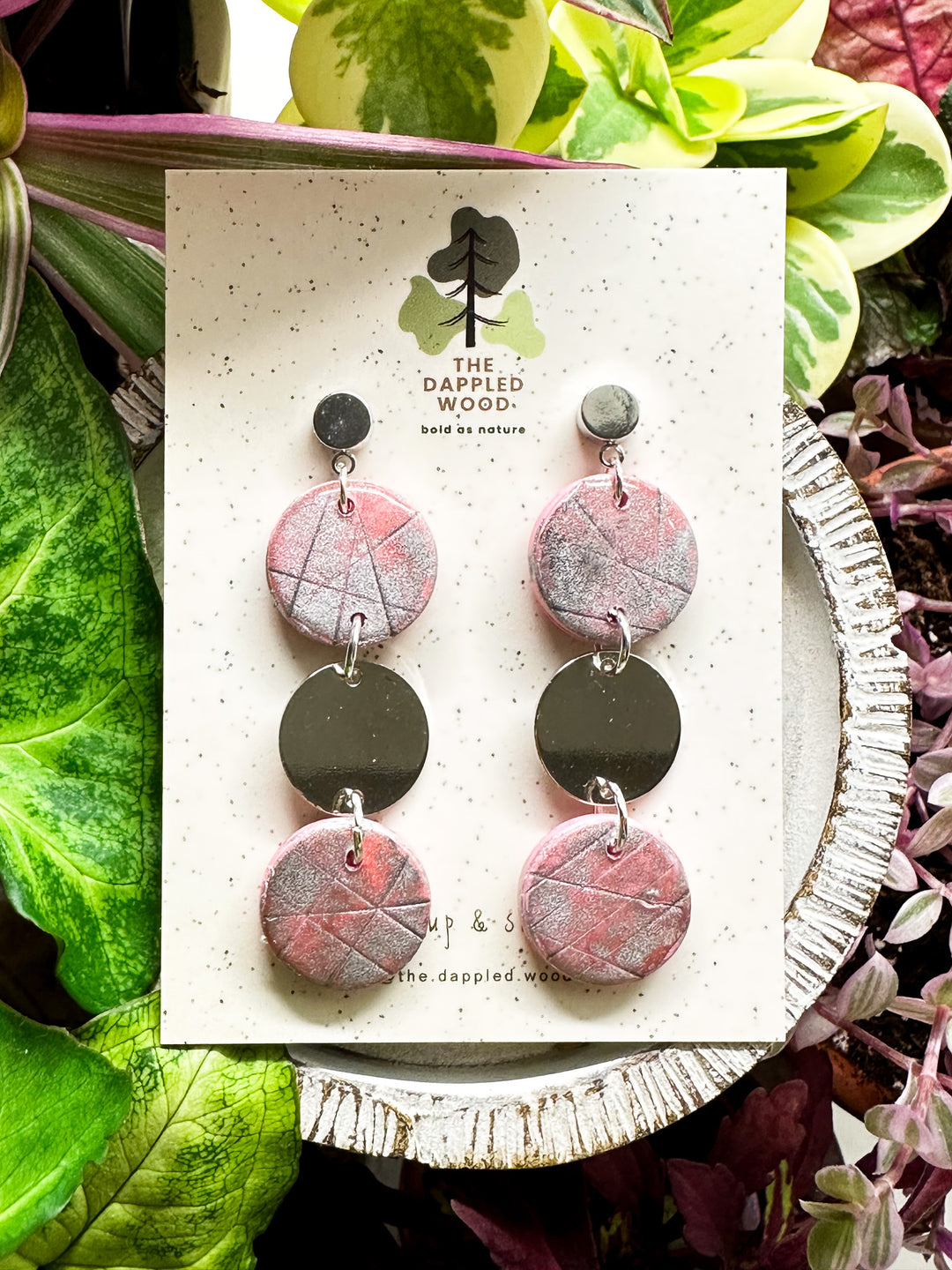 Handcrafted pink polymer clay disk earrings with silver shimmer and a smooth silver connector charm, displayed on a speckled card with The Dappled Wood logo against a lush backdrop of green plants.