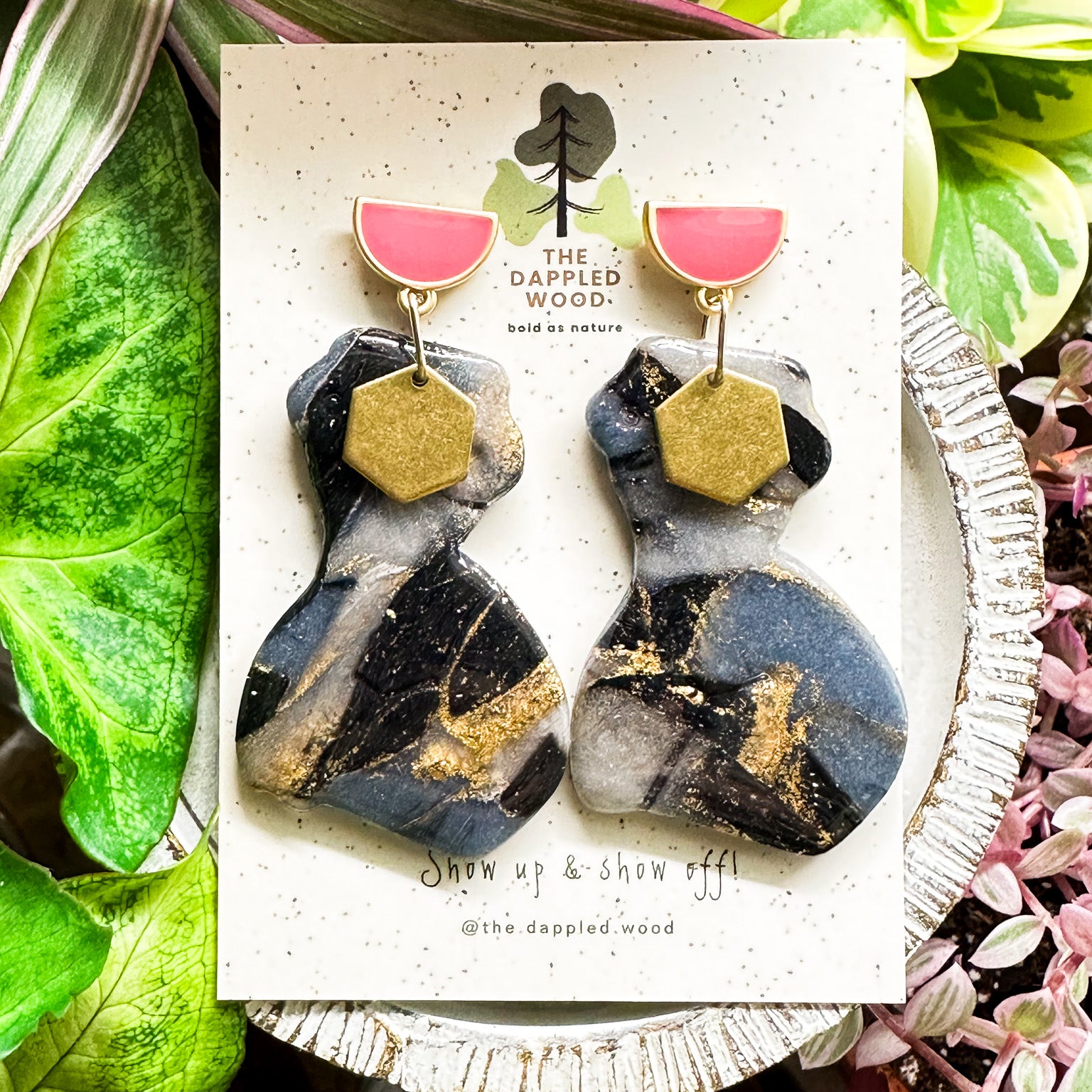 A pair of polymer clay earrings displayed on a speckled card. The earrings feature a marbled black and gray pattern with shimmering gold accents and are in the shape of a curvy woman that is adorned with a brass hexagon charm. The earrings hang from pink and gold half moon posts. They are surrounded by vibrant green and pink plants. The card reads 'The Dappled Wood: bold as nature' and 'Show up & show off!' with the handle '@the.dappled.wood'