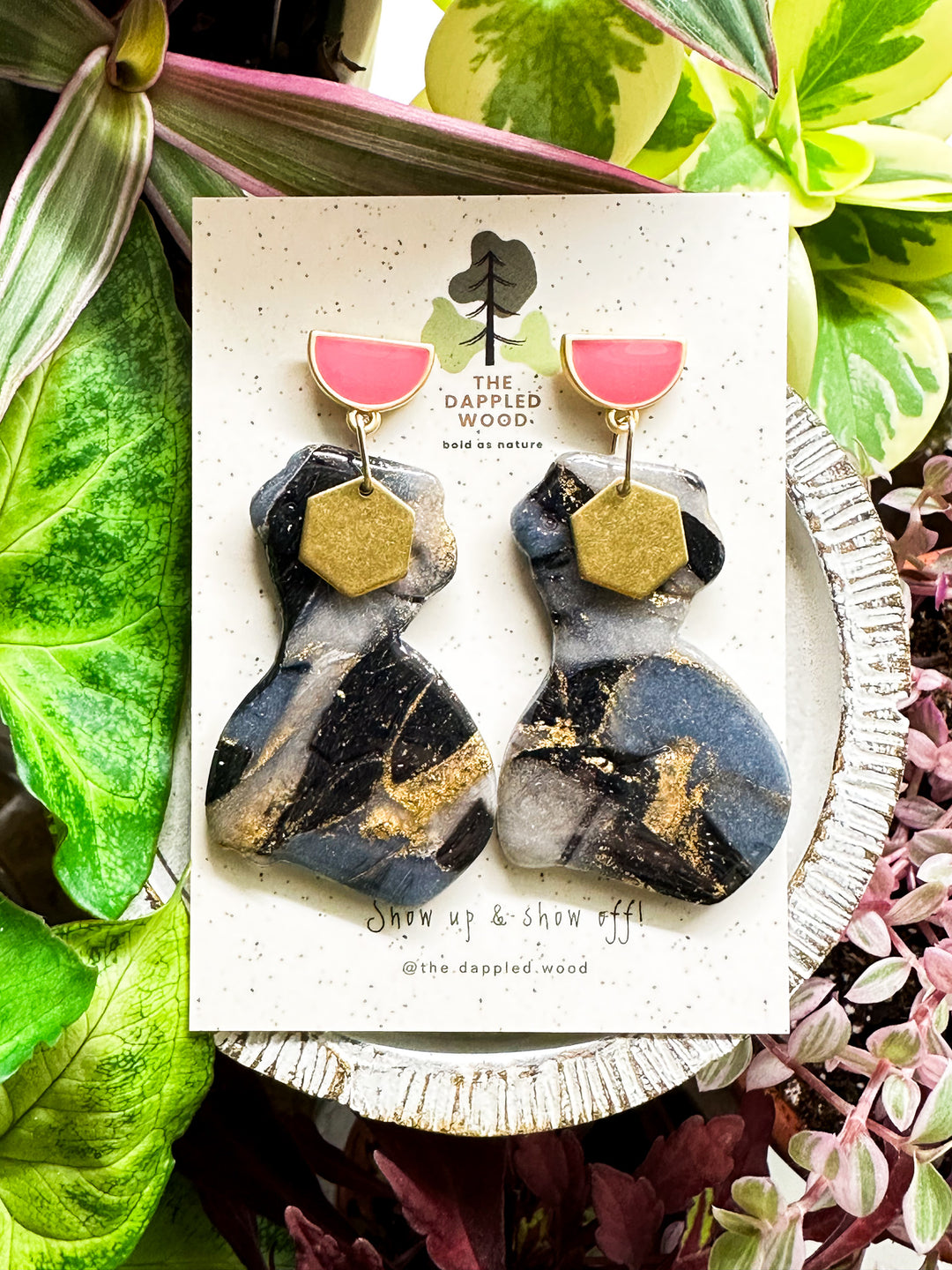 A pair of polymer clay earrings displayed on a speckled card. The earrings feature a marbled black and gray pattern with shimmering gold accents and are in the shape of a curvy woman that is adorned with a brass hexagon charm. The earrings hang from pink and gold half moon posts. They are surrounded by vibrant green and pink plants. The card reads 'The Dappled Wood: bold as nature' and 'Show up & show off!' with the handle '@the.dappled.wood'