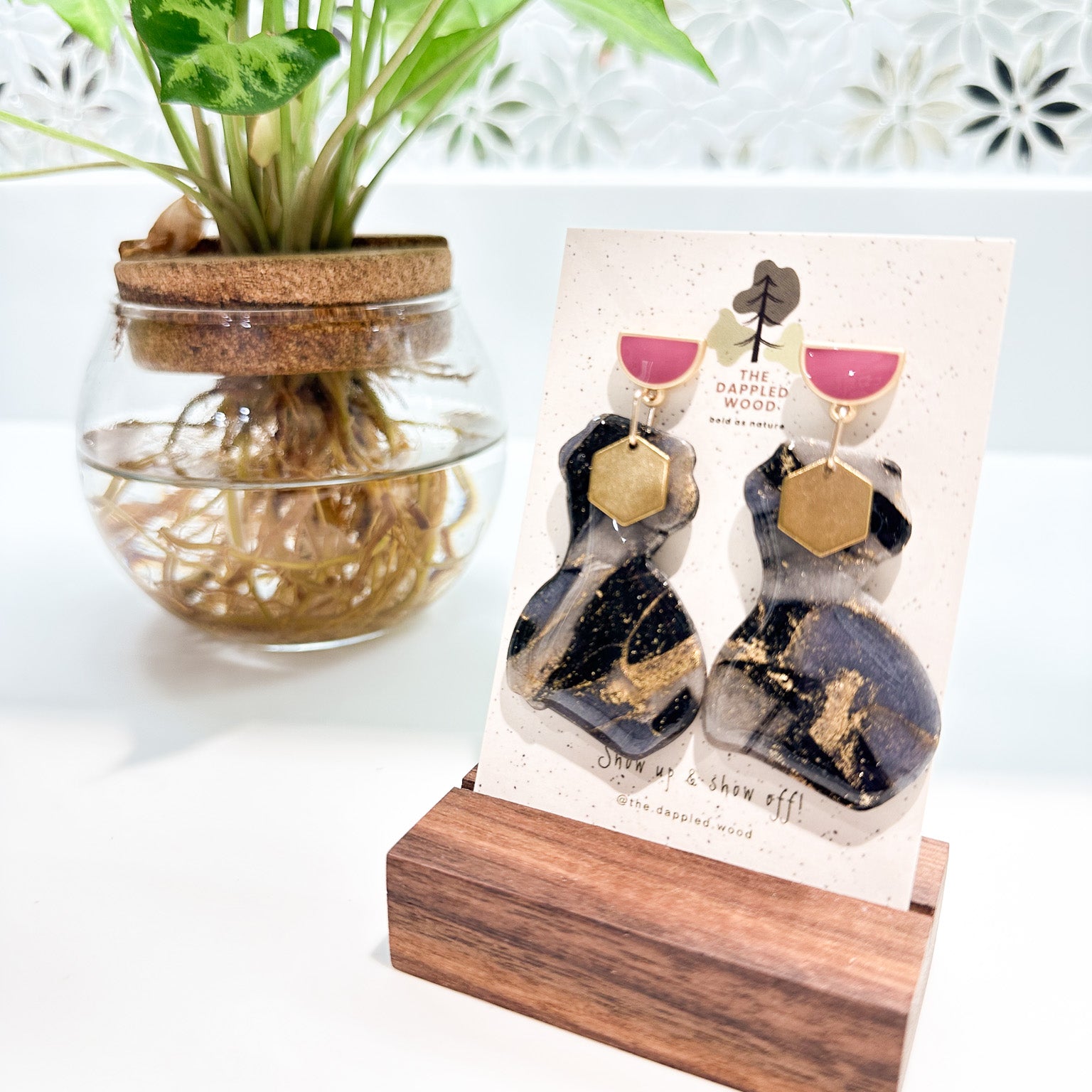 A pair of polymer clay earrings displayed on a speckled card with The Dappled Wood logo. The earrings feature a marbled black and gray pattern with shimmering gold accents and are in the shape of a curvy woman that is adorned with a brass hexagon charm. The earrings hang from pink and gold half moon posts. They are flanked by a green plant in a clear vase where roots are shown growing in water with a floral tile mosaic in the background. 