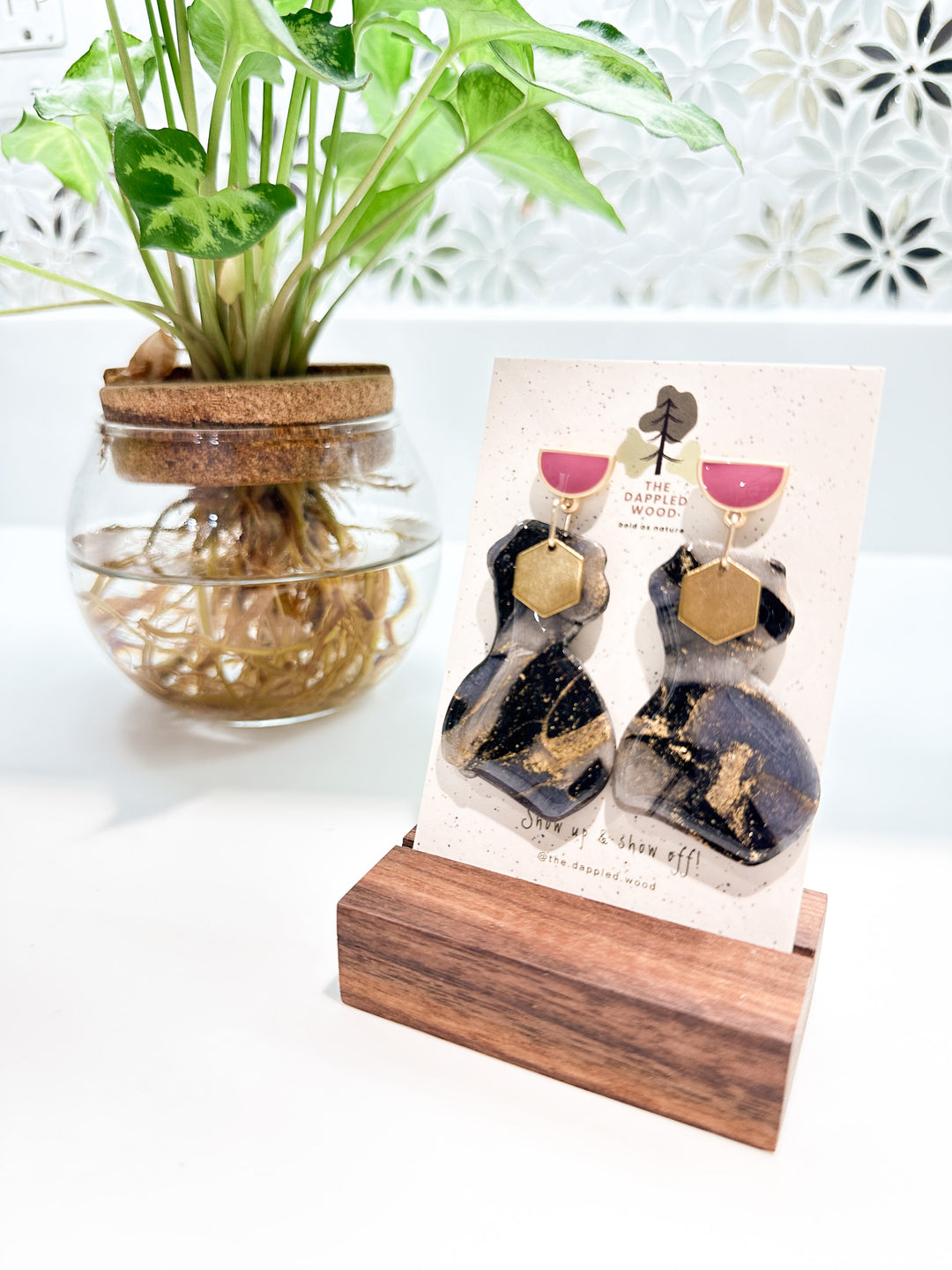 A pair of polymer clay earrings displayed on a speckled card with The Dappled Wood logo. The earrings feature a marbled black and gray pattern with shimmering gold accents and are in the shape of a curvy woman that is adorned with a brass hexagon charm. The earrings hang from pink and gold half moon posts. They are flanked by a green plant in a clear vase where roots are shown growing in water with a floral tile mosaic in the background. 