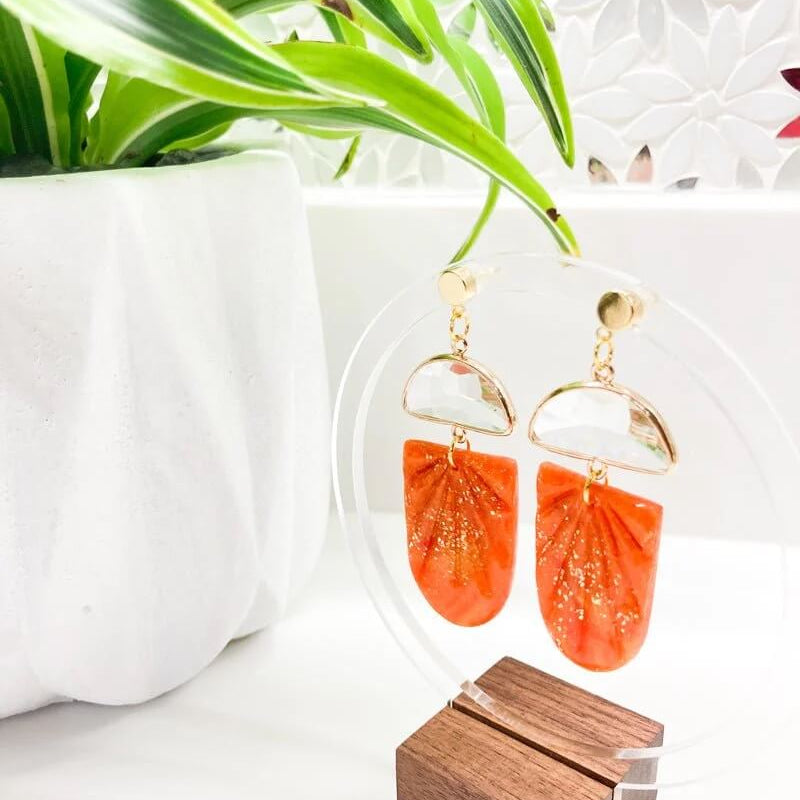 An elegant pair of handcrafted orange polymer clay earrings with a sparkling glass gem, displayed against a white background with a lush green plant and a wooden stand.