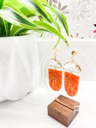 An elegant pair of handcrafted orange polymer clay earrings with a sparkling glass gem, displayed against a white background with a lush green plant and a wooden stand.