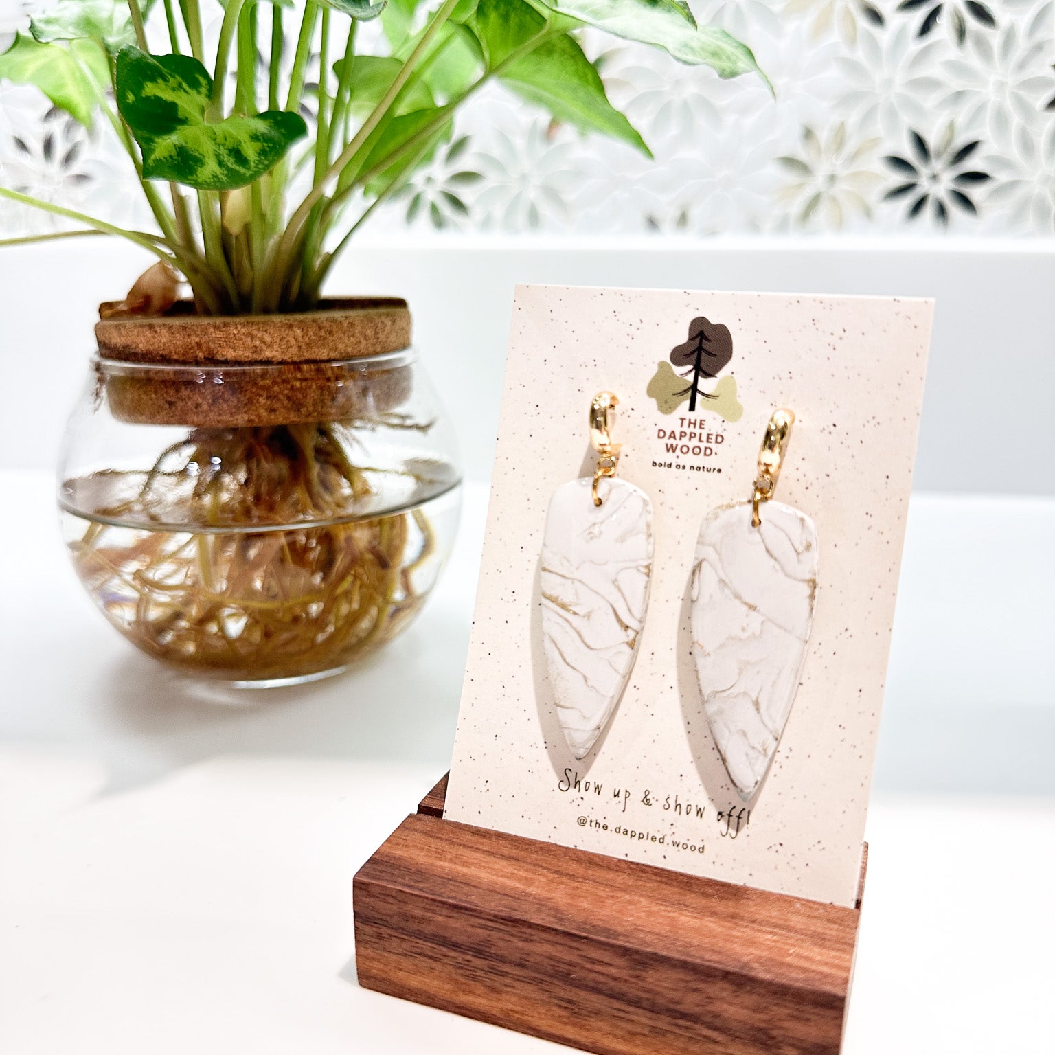 Handcrafted white marble-textured dagger earrings with gold hoops from The Dappled Wood displayed on a wooden stand with a green plant backdrop.