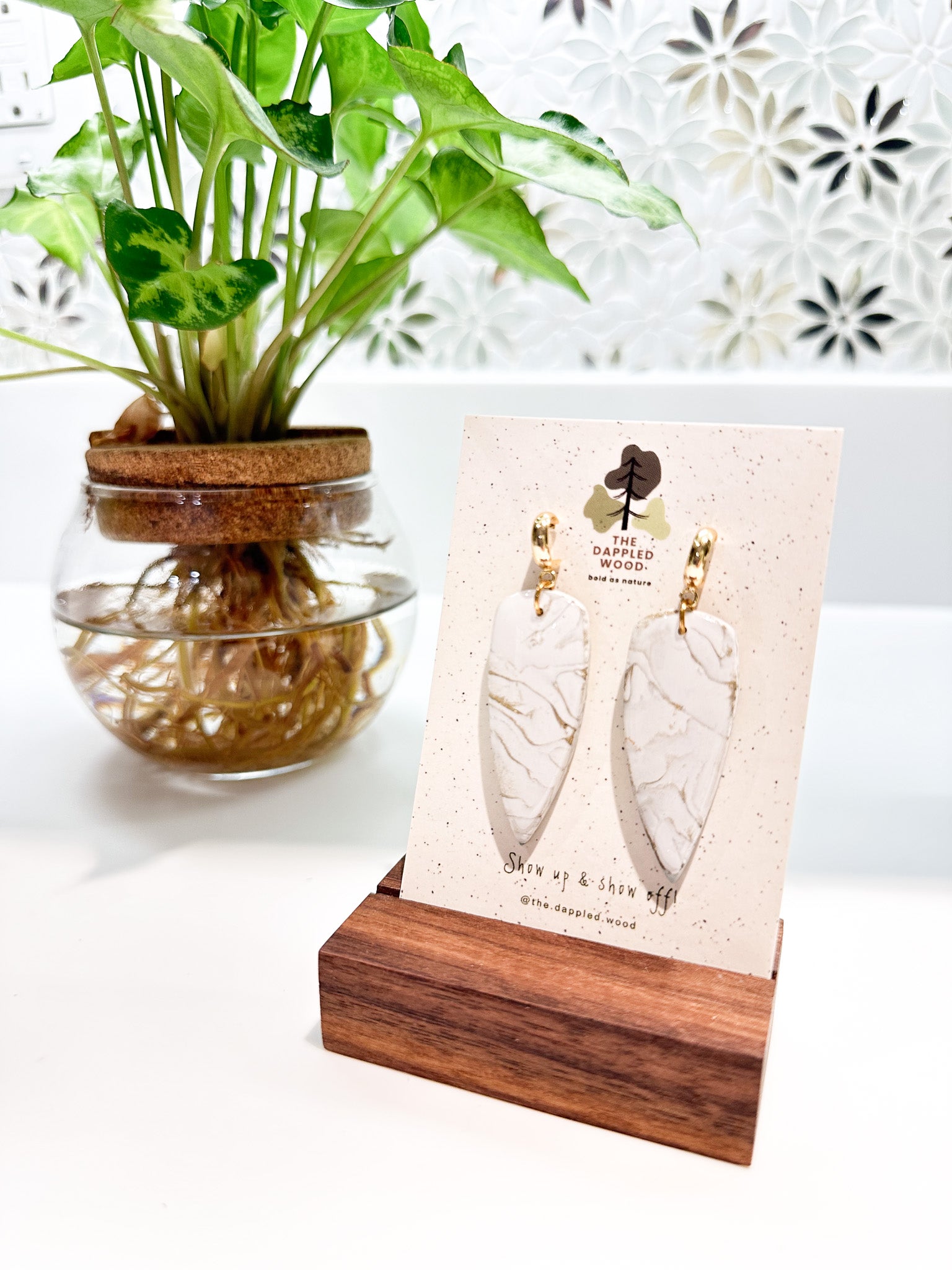 Handcrafted white marble-textured dagger earrings with gold hoops from The Dappled Wood displayed on a wooden stand with a green plant backdrop.