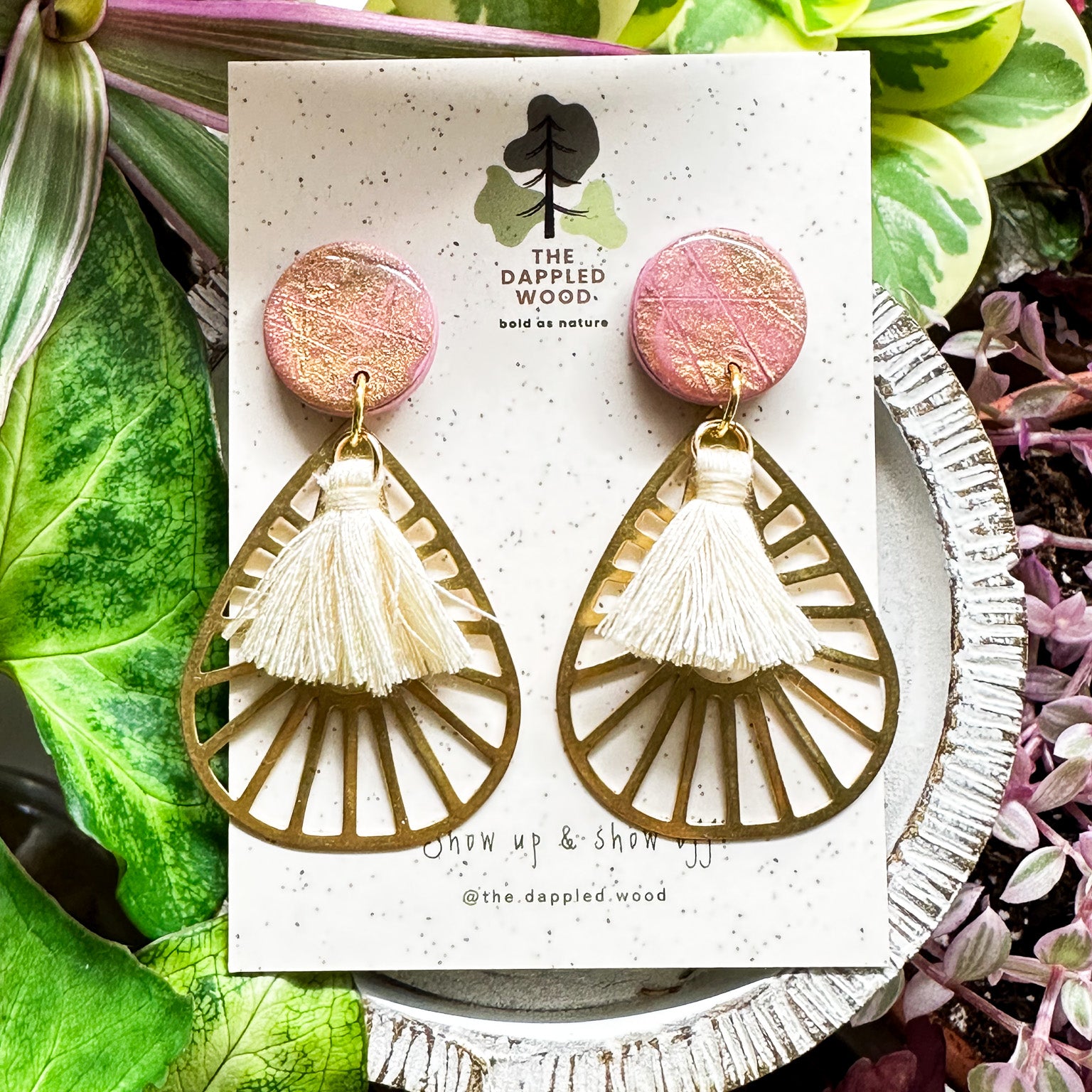 Handcrafted polymer clay earrings named 'Willa', showcasing a sparkling pink circular top with an openwork gold fan design and delicate tassels, presented against a speckled card with The Dappled Wood logo, surrounded by vibrant green and pink plants.