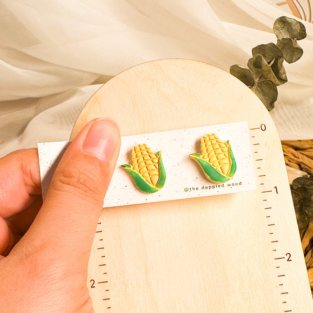 Yellow corn stud earrings with green husks on an earring card beside an inch ruler