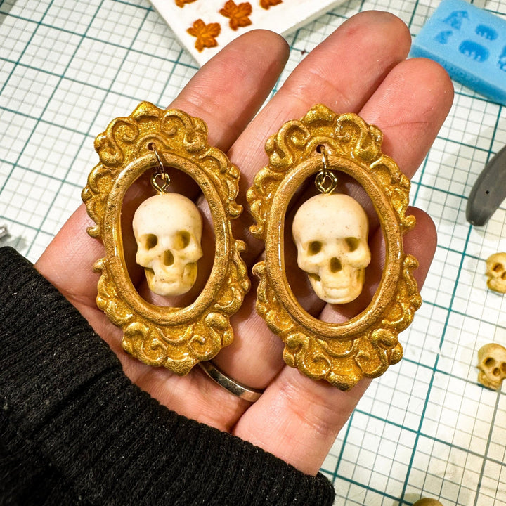 Drusilla ivory skull earrings in ornate gold frames held in a hand.