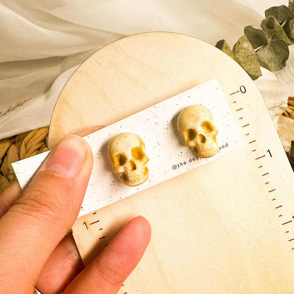 Cream Sybil 3D skull stud earrings on an earring card beside an inch ruler