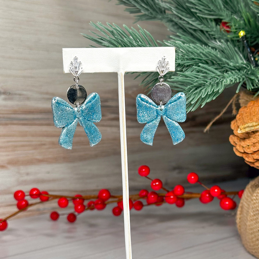 The Dappled Wood Ho Ho Bow earrings in Frost Blue, a winter-inspired icy hue with a delicate shimmer. A festive yet elegant earring choice for the holiday season.