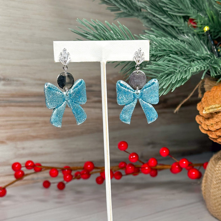 The Dappled Wood Ho Ho Bow earrings in Frost Blue, a winter-inspired icy hue with a delicate shimmer. A festive yet elegant earring choice for the holiday season.