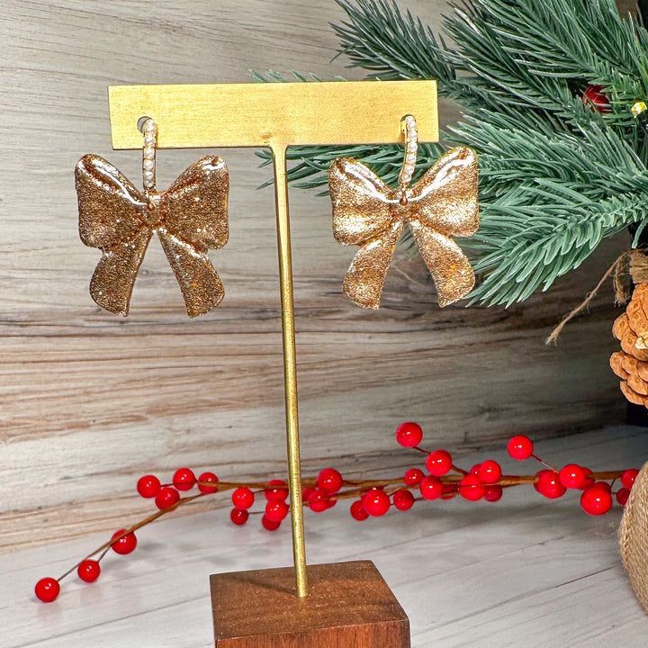 The Dappled Wood Ho Ho Bows earrings in Gold, shimmering with a warm metallic finish. These handcrafted polymer clay bows add a touch of luxury to any holiday outfit.