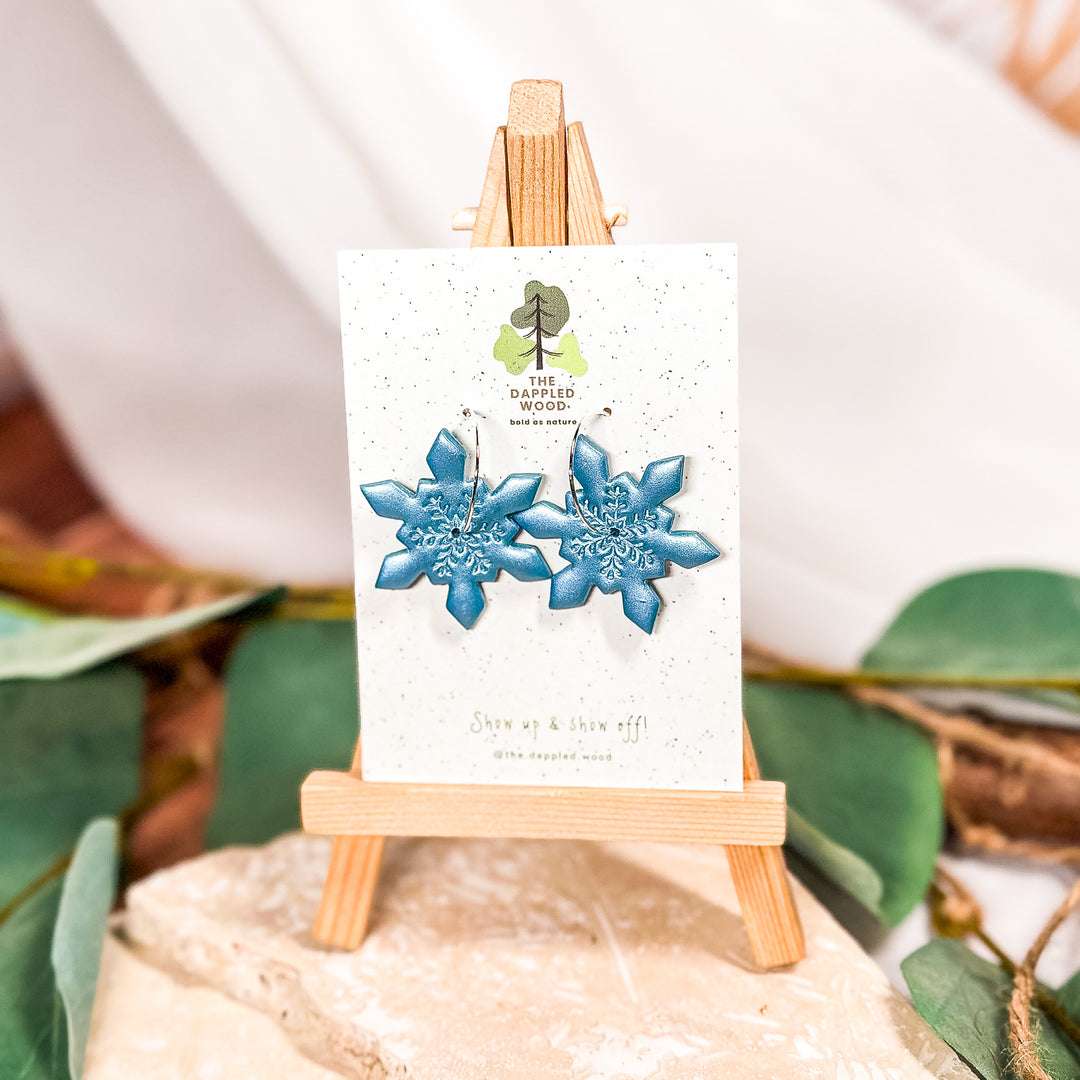 The Dappled Wood Holly Jolly Vibes snowflake earrings from the Holidays 2024 collection featuring a winter-inspired polymer clay design and surgical steel hoops, suitable for sensitive ears.